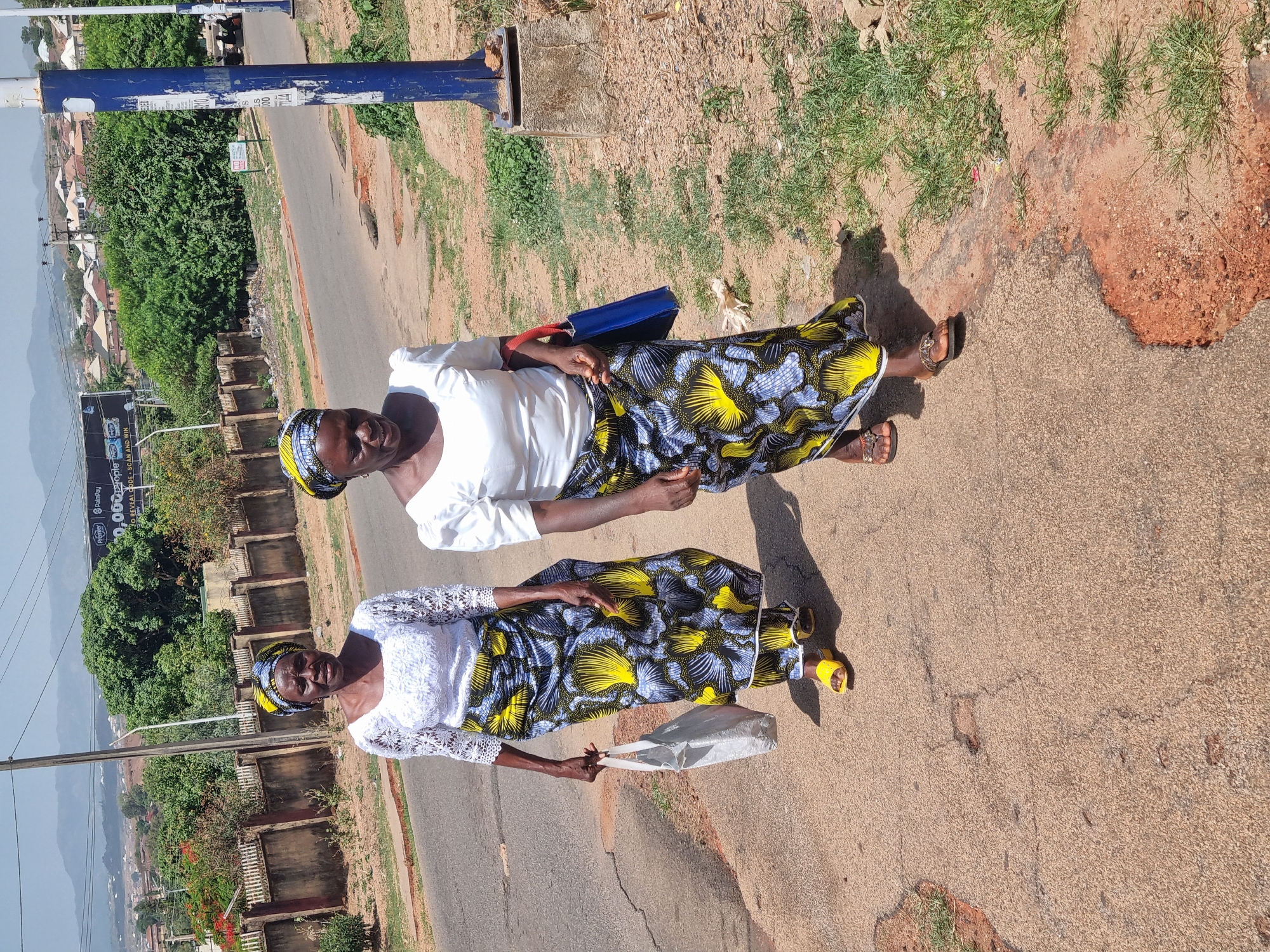 Two middle-aged African women stand on a paved street in Jos, Nigeria. The woman on the left wears a white lace top, a black and yellow patterned skirt, yellow sandals, and a matching head wrap, holding a white plastic shopping bag. The woman on the right wears a white long-sleeved shirt, a similar patterned skirt, and brown sandals, with a matching head wrap, and carries a blue purse over her shoulder. They appear to be posing. The street is bordered by a sidewalk with patchy grass and reddish soil. A blue metal pole and a billboard are visible in the background, along with buildings and greenery, including trees. In the distance, hills can be seen under a clear sky. The street shows signs of wear and tear, with cracks and patches.