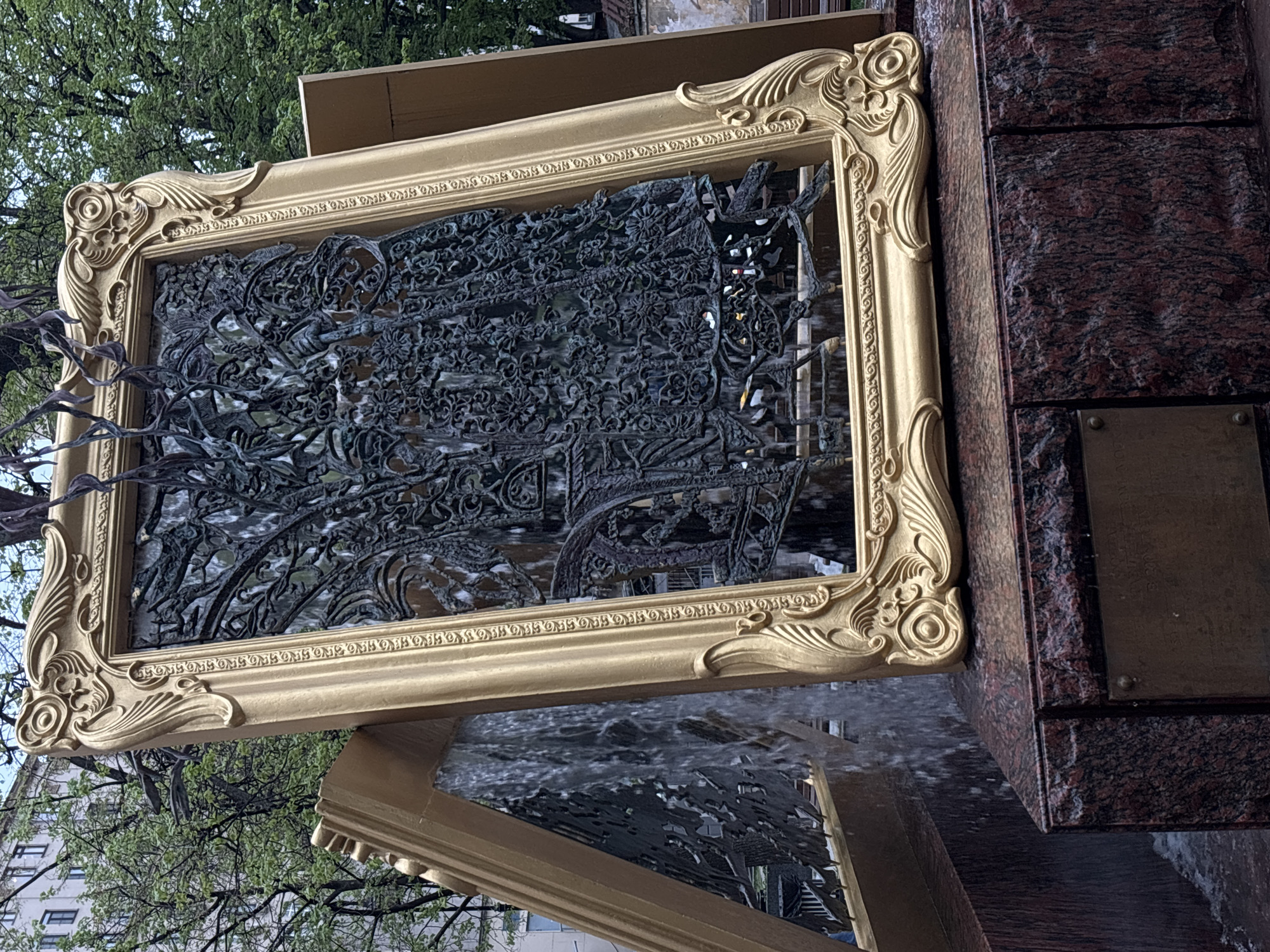 The image shows a metallic structure with intricate patterns encased in a golden frame, appearing to be part of a fountain. Water flows from the structure. The frame is attached to a stone structure. Part of the structure reflects in the water. Trees and buildings are visible in the background. Based on the style of the structure, it appears to be in the city of Moscow, Russia.