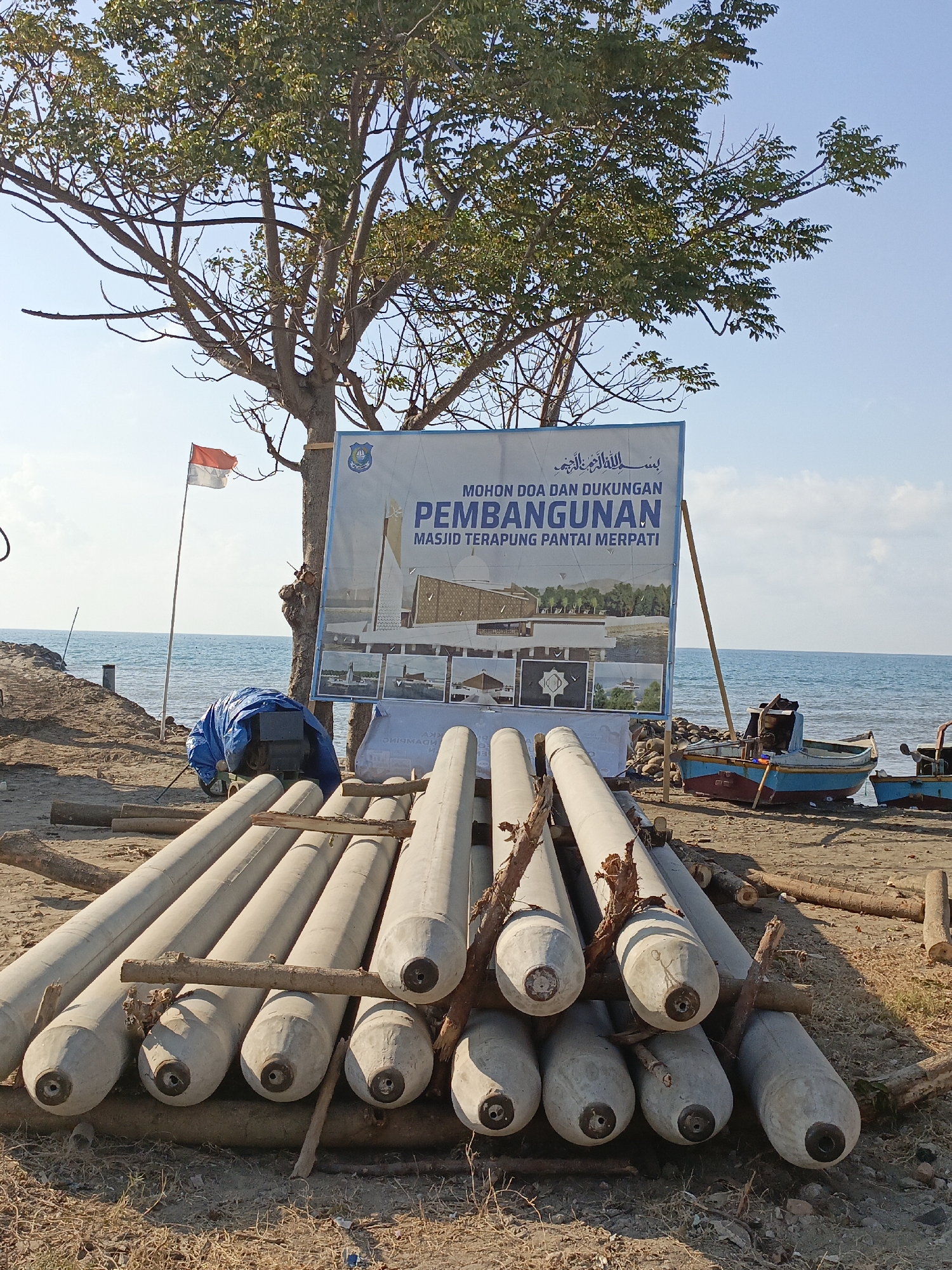 An outdoor daytime scene shows a beach area with construction materials, a sign, and the sea in the background. In the foreground, a pile of large, cylindrical concrete structures, possibly pilings or pipes, are arranged horizontally on the ground, supported by wooden beams. To the left of the concrete structures, a blue tarp covers an object that appears to be machinery. Further left, an Indonesian flag waves from a flagpole.
Behind the construction materials and tarp, a large sign is propped up by a wooden pole. The sign is blue and white and features text in Arabic and Indonesian, along with images depicting a mosque. The Indonesian text reads "MOHON DOA DAN DUKUNGAN PEMBANGUNAN MASJID TERAPUNG PANTAI MERPATI" which translates to "PRAYER AND SUPPORT FOR THE CONSTRUCTION OF THE FLOATING MOSQUE AT MERPATI BEACH."
The background consists of a sandy beach, leading to the sea. Two small boats are visible on the beach near the water's edge. The sea is calm and extends to the horizon, meeting a clear blue sky. A large tree with green foliage stands behind the sign, its branches extending over the scene. The location is identified as Pantai Merpati in Bulukumba, Indonesia, a coastal area known for its fishing communities and developing infrastructure. The overall atmosphere suggests an ongoing construction project in a coastal environment.
