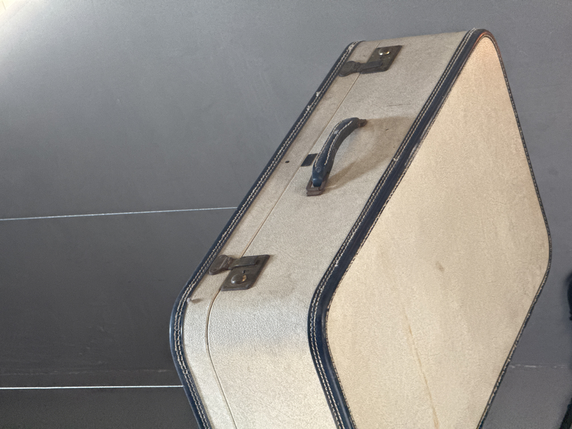 A vintage-style suitcase stands upright, positioned diagonally against a plain, dark gray background. The suitcase features a light beige or cream-colored body with a slightly textured surface, outlined by a dark blue or black trim secured with contrasting white stitching. A black, textured handle is centrally located on the top edge, and two metallic clasps, appearing somewhat aged, are visible further along the top.
The suitcase has a distinctive shape, with rounded corners and a unique profile that widens from the top edge downwards, giving it an elongated hexagonal or trapezoidal appearance when viewed from its end. Subtle signs of wear and age are visible on the suitcase's surface and hardware, contributing to its vintage aesthetic. The background is simple and monochromatic, with faint horizontal lines that might indicate shelves or panels. No specific activity is depicted, and there is no visible text. The image was captured in Apetatitlán, Mexico.