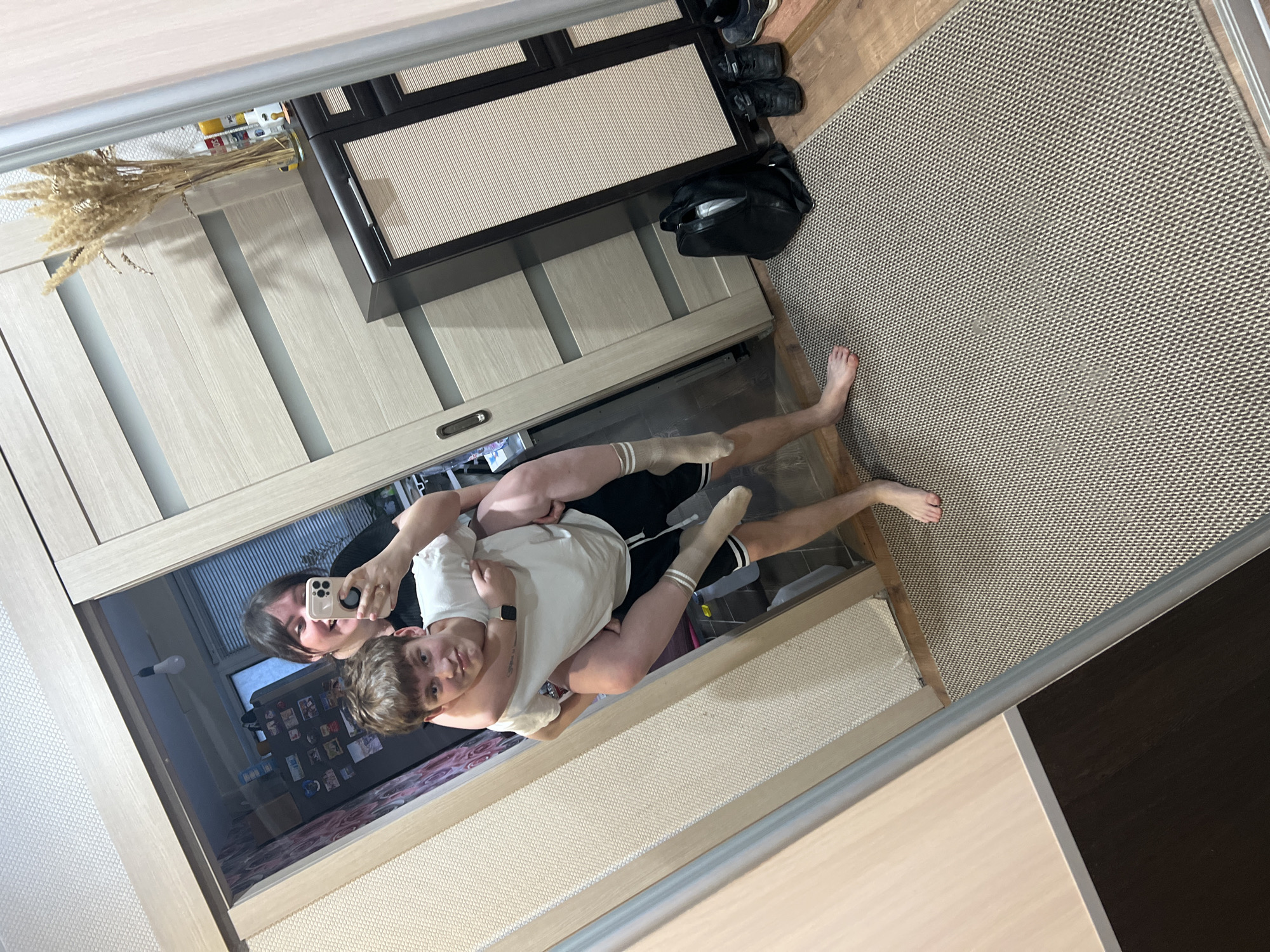 The image captures a mirror reflection of an indoor domestic scene. Two individuals are present: a female, visible from the chest up, with dark hair, smiling and holding a light-colored smartphone to capture the image. She wears a black top and a ring on her left hand. She is giving a piggyback ride to a male youth with light brown, messy hair, who looks towards the mirror with a slight smirk. He wears a white t-shirt and black shorts with white side stripes. His left foot is bare, while his right leg wears a light-colored, striped knee-high sock. A dark wristband is on his left wrist.
The environment features light-colored textured wallpaper and a wooden floor, partially covered by a light brown woven rug. A light wood-toned sliding door or wardrobe with horizontal frosted panels is visible on the left. To the right, a dark brown cabinet with slatted doors displays a glass vase containing dried ornamental grass and several aerosol cans. A black backpack and multiple pairs of dark shoes are situated on the floor adjacent to the cabinet. Through an open doorway reflected in the background, a room is visible, containing a window with horizontal blinds, a refrigerator adorned with magnets, and a bare incandescent lightbulb suspended from the ceiling. The wall in the background room features patterned wallpaper. The scene depicts a typical interior of a residence.