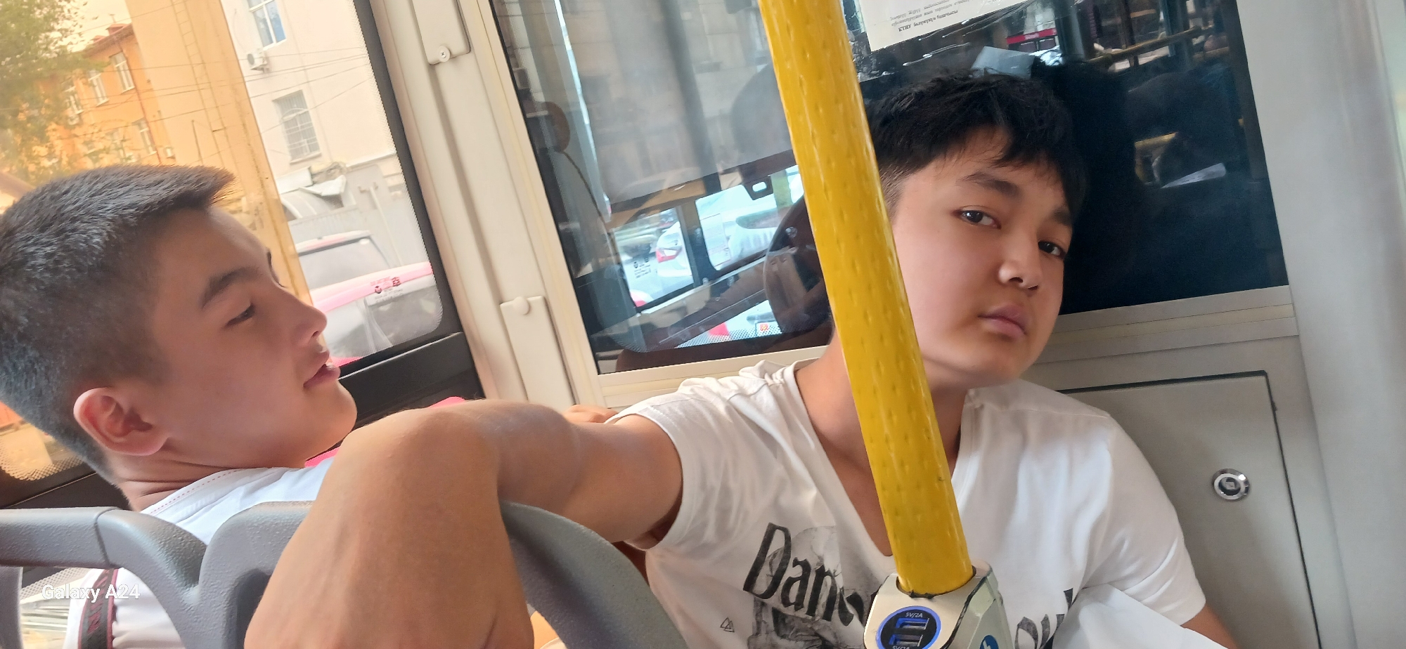 Two young boys are seated on a bus. The boy on the left, facing away from the camera, has short dark hair and is wearing a white t-shirt. He is looking out the window and appears to be speaking. The boy on the right, facing the camera, also has short dark hair and is wearing a white t-shirt with a graphic design. He is looking directly at the camera with a neutral expression. Between them is a yellow pole, a common feature on public transportation. The bus interior is visible, with windows showing buildings and vehicles outside, suggesting an urban environment. The overall scene is consistent with being in Bishkek City, Kyrgyzstan, where such public transportation and urban landscapes are common.