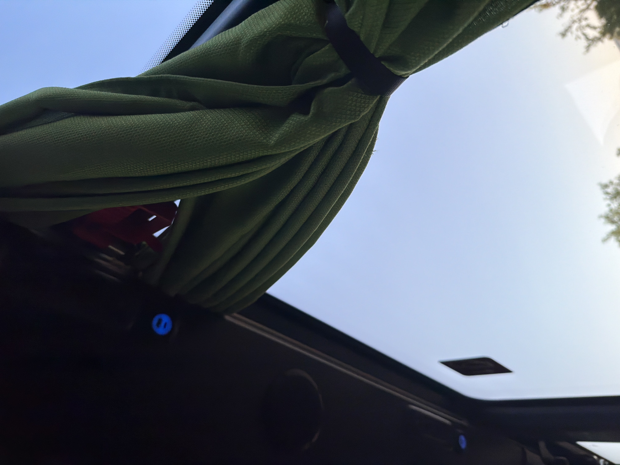 The image shows a close-up view of a green curtain inside a vehicle, likely a bus or van, as suggested by the dark interior paneling and the view of the sky through a window. The curtain is draped and secured with a black band. A small, circular blue light is visible on the dark interior panel to the left. The sky outside is a clear, pale blue, indicating it is daytime. The location of Shtarkovo, Bulgaria is not depicted in this image.