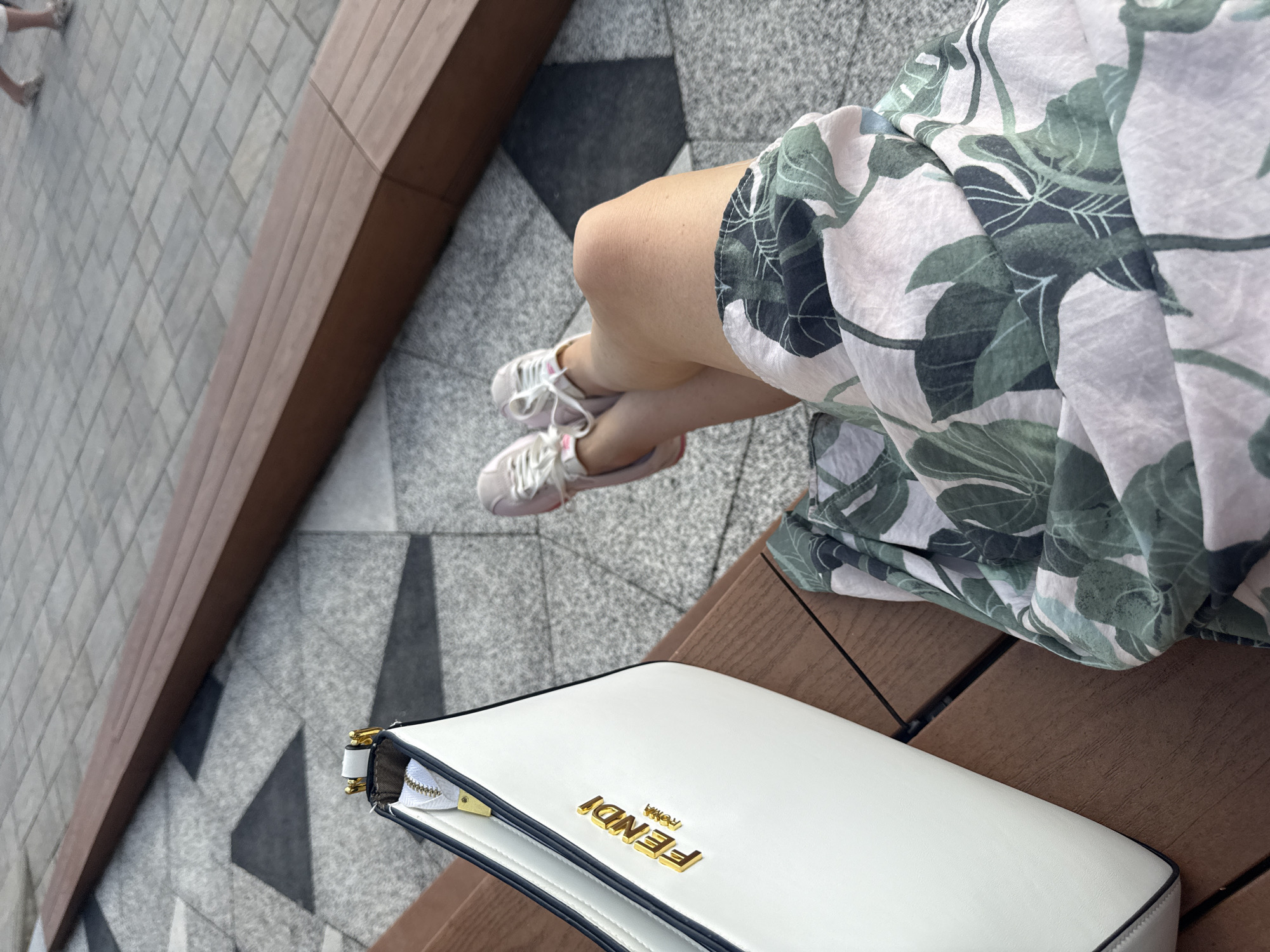 The image shows a close-up of a person's legs and a white Fendi bag, placed on a wooden bench. The person is wearing a light-colored dress with a green leaf print and white sneakers. The background features a patterned stone pavement. The scene suggests a casual outing, possibly in a park or an outdoor seating area. The lighting indicates it's daytime. The bag has the "FENDI ROMA" logo in gold lettering. The location context is Sochi, Russia.