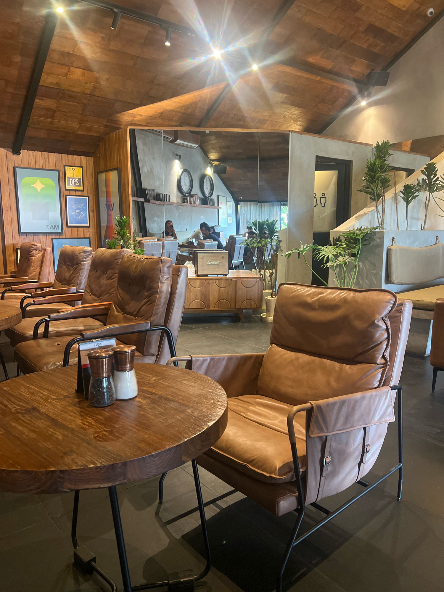 The image depicts the interior of a modern cafe with a warm, earthy ambiance. The ceiling is clad in wooden planks, creating a rustic effect, and is illuminated by track lighting that casts bright spotlights. On the left wall, several framed art prints are displayed, adding a touch of color and personality to the space.
The seating area features several comfortable-looking brown leather armchairs arranged around round wooden tables. In the foreground, a close-up of one of these tables reveals a salt and pepper shaker set. The chairs have a contemporary design with metal frames and leather upholstery.
In the background, a large mirror reflects a portion of the cafe, showing more seating and a person sitting at a table. The cafe has a concrete wall section where a set of clocks is visible, and to the right, a doorway is marked with male and female restroom symbols. Plants are strategically placed throughout the cafe, adding a touch of greenery. The flooring is dark and reflective. This scene is located in North Kuta, Indonesia.