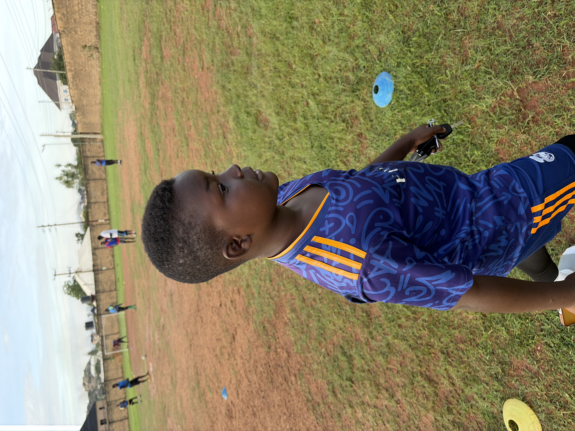 The image captures an outdoor scene on a partially grassy, reddish-brown dirt field in Jos, Nigeria. In the foreground, a young child with dark skin and short hair is visible in profile, facing right. The child wears a navy blue athletic jersey featuring an abstract lighter blue pattern, an orange V-neck collar, and three orange stripes on each shoulder. Matching navy blue shorts with orange side stripes are also worn. A set of silver keys with a black fob is held in the child's right hand. A yellow training cone is positioned on the ground in the lower-left foreground, and a blue training cone is in the lower-right mid-ground.
In the mid-ground, a soccer training session is underway with approximately six to eight individuals, primarily young adults, scattered across the field. They are dressed in various colored athletic attire, including blue, red, and white shirts. A white soccer goal with a net is visible towards the left side of the mid-ground, and a white soccer ball rests on the ground near the center. Another blue training cone is visible further back on the left.
The background features a low, light-brown perimeter wall extending horizontally across the scene. Beyond this wall, various residential buildings are visible, including a prominent structure with a dark, pyramid-shaped roof. Utility poles and overhead power lines are also present. Distant hills or mountains are discernible under a pale, cloudy sky, indicating daytime conditions.