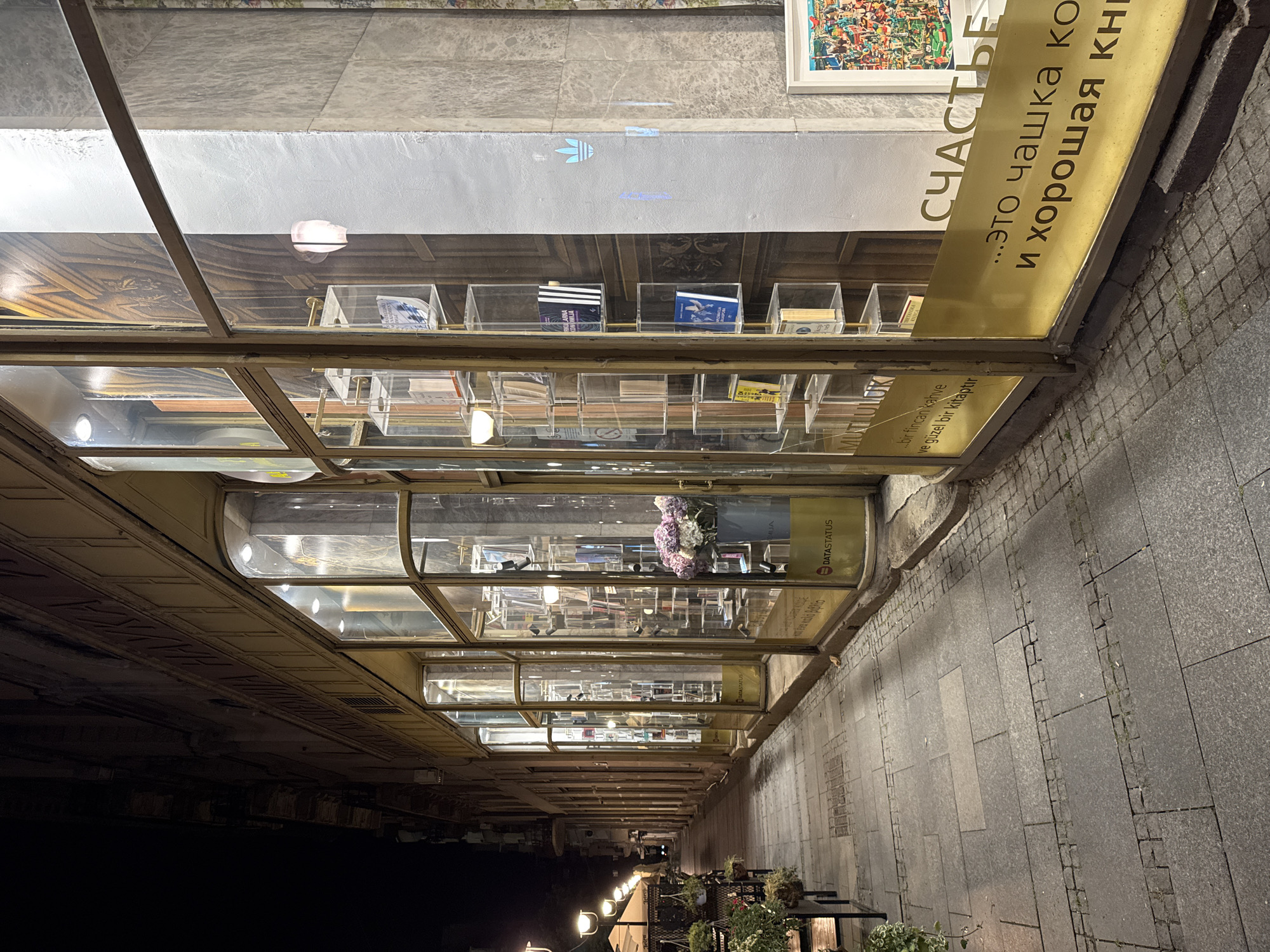 The image shows the exterior of a bookstore in Belgrade, Serbia, likely at night. The bookstore has a series of glass-enclosed display cases running along the sidewalk. The cases contain books, and some have decorative elements like flowers. Signs in Turkish and Russian with the words "happiness", "coffee", and "book" are visible. The sidewalk is paved with grey stone and there are some plants near the bookstore. The image is taken from a low angle.