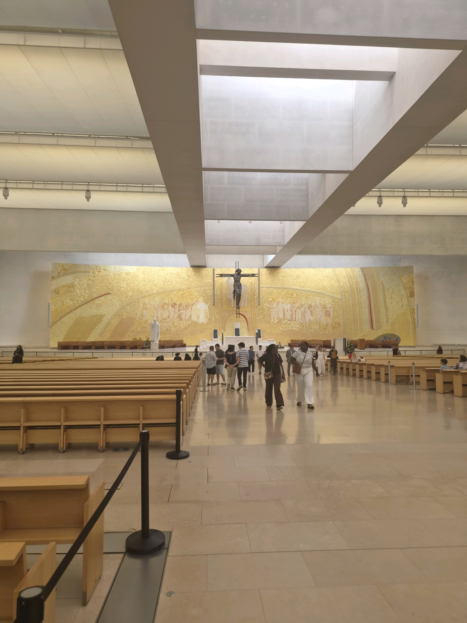 The image captures a wide-angle view of the interior of the Sanctuary of Fátima in Fátima, Portugal. The vast space features rows of light-colored wooden pews arranged in sections. The floor is paved with large, light beige tiles. A prominent feature is a large, golden mosaic mural on the far wall behind the altar area, depicting religious figures and scenes. Above the mosaic, a large crucifix with a dark figure of Christ is visible, along with a white statue of a religious figure to the left of the altar.
Several people are present, walking and standing in the central aisle and near the pews. They are casually dressed, suggesting they are visitors or worshippers. Two women in the foreground are walking towards the camera, one wearing white pants and a striped top, and the other in dark pants and a dark top. Other individuals are seen in smaller groups further in the background.
The ceiling is a modern design with large, recessed lighting panels and prominent horizontal beams, giving the space an airy and contemporary feel. Natural light appears to be filtering in from skylights or high windows. The overall impression is one of a large, modern, and serene religious space.
