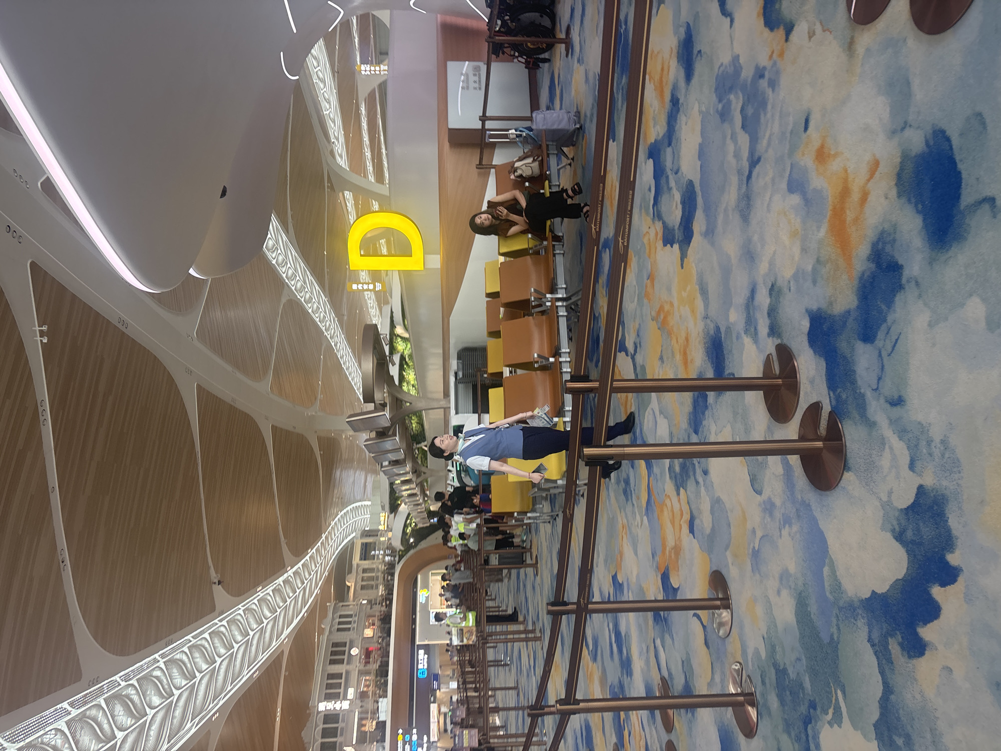 The image depicts an interior view of a modern airport terminal, likely located in Baiyun District, China, as suggested by subtle signage elements. The scene features a brightly lit, spacious area with a high ceiling exhibiting an intricate, organic design with wooden panels and white curved elements. A prominent yellow letter "D" is displayed on a wall, indicating a gate or zone.
In the foreground, several stanchions with retractable belts guide a pathway across a carpeted floor adorned with a vibrant, abstract pattern resembling clouds with blue and orange hues. A woman in a blue uniform, possibly an airport employee, walks through the frame, holding a piece of paper and a phone. She is looking to her right, and appears to be in motion.
Further back, rows of empty brown and yellow seating are arranged in a waiting area. A woman is seated on one of the chairs, looking down at her phone. Luggage, including a suitcase and a backpack, is visible near her. Other individuals, including airport staff in high-visibility vests, are scattered throughout the background, some standing near check-in counters or in queues. The overall environment suggests a functional and aesthetically designed public transit hub.