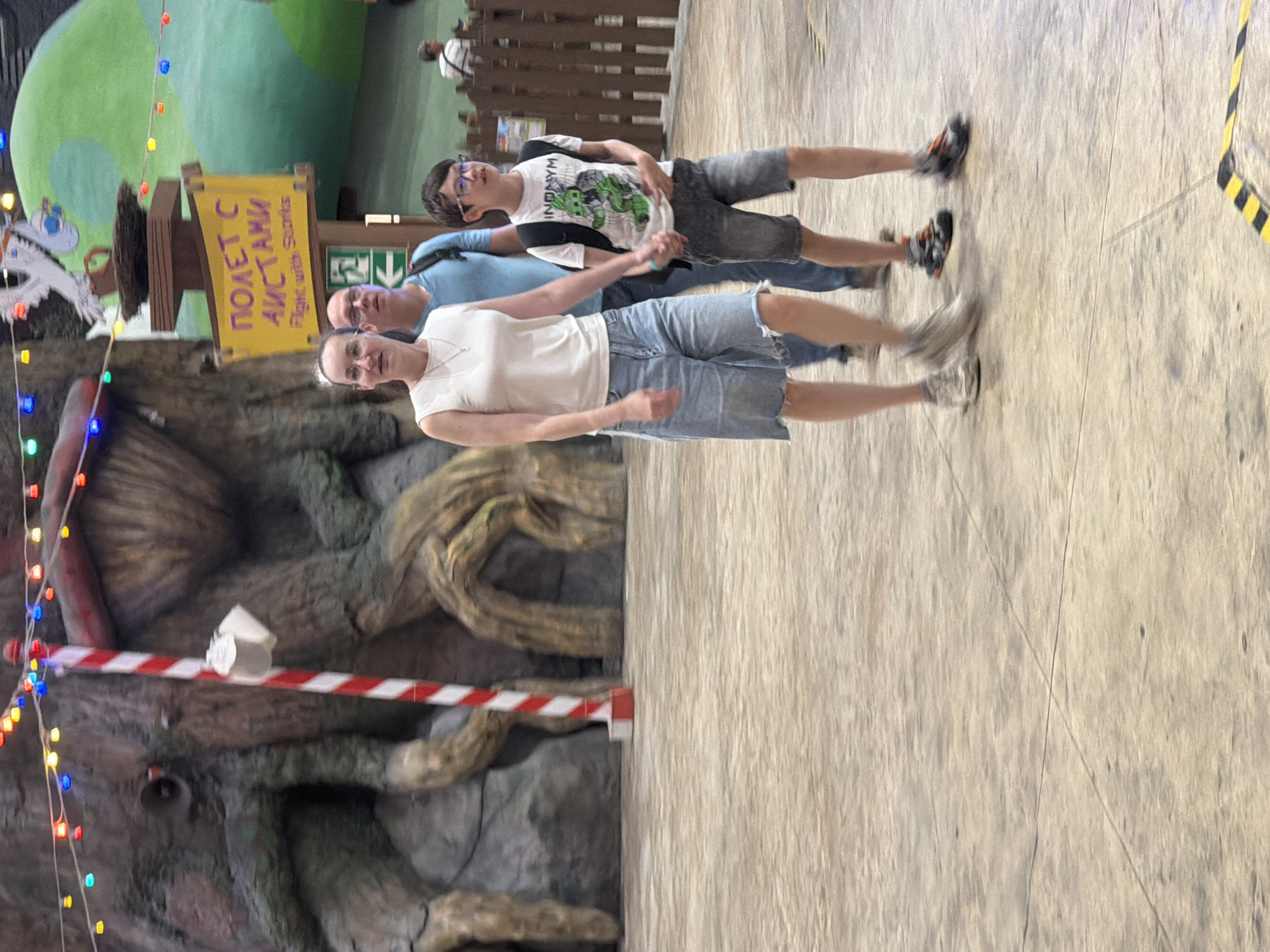 In an indoor amusement park or themed attraction, a family of three walks together. The mother, in the foreground, wears a white sleeveless shirt and denim shorts, her left arm linked with her son's. Her son, on her right, wears a white t-shirt with a graphic print and dark shorts, his left hand holding his mother's and his right hand holding a small object. Behind them, the father, in a blue t-shirt and jeans, walks with his arm extended. The background features a large, tree-like structure and a sign with Cyrillic text and "Flight with Storks." Colored lights are strung across the upper portion of the scene. The flooring is a light-colored, textured material with visible seams.