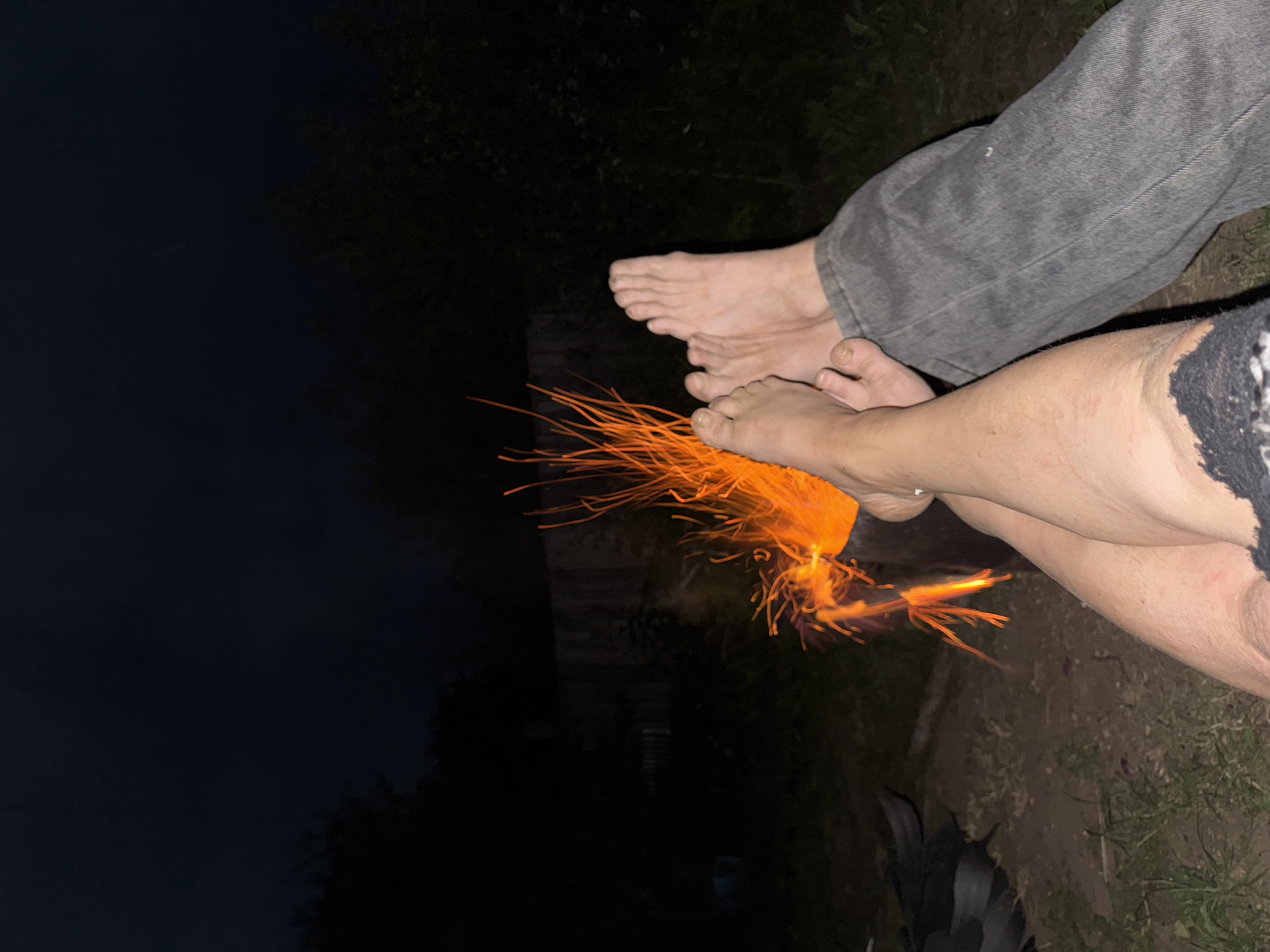 Two individuals, one wearing grey denim jeans and the other a black lace-trimmed garment, sit outdoors at night. Their bare feet are visible in the foreground, with one set of feet positioned closer to the camera and slightly lower than the other. A brightly burning sparkler, emitting a shower of orange sparks, is held between their feet, illuminating the immediate area with its warm glow. The background is dark, suggesting nighttime, and appears to be an outdoor setting with some faint indications of foliage and structures. The overall mood is relaxed and convivial, as the subjects appear to be enjoying a festive activity. The time of day is clearly night. There is no visible text.