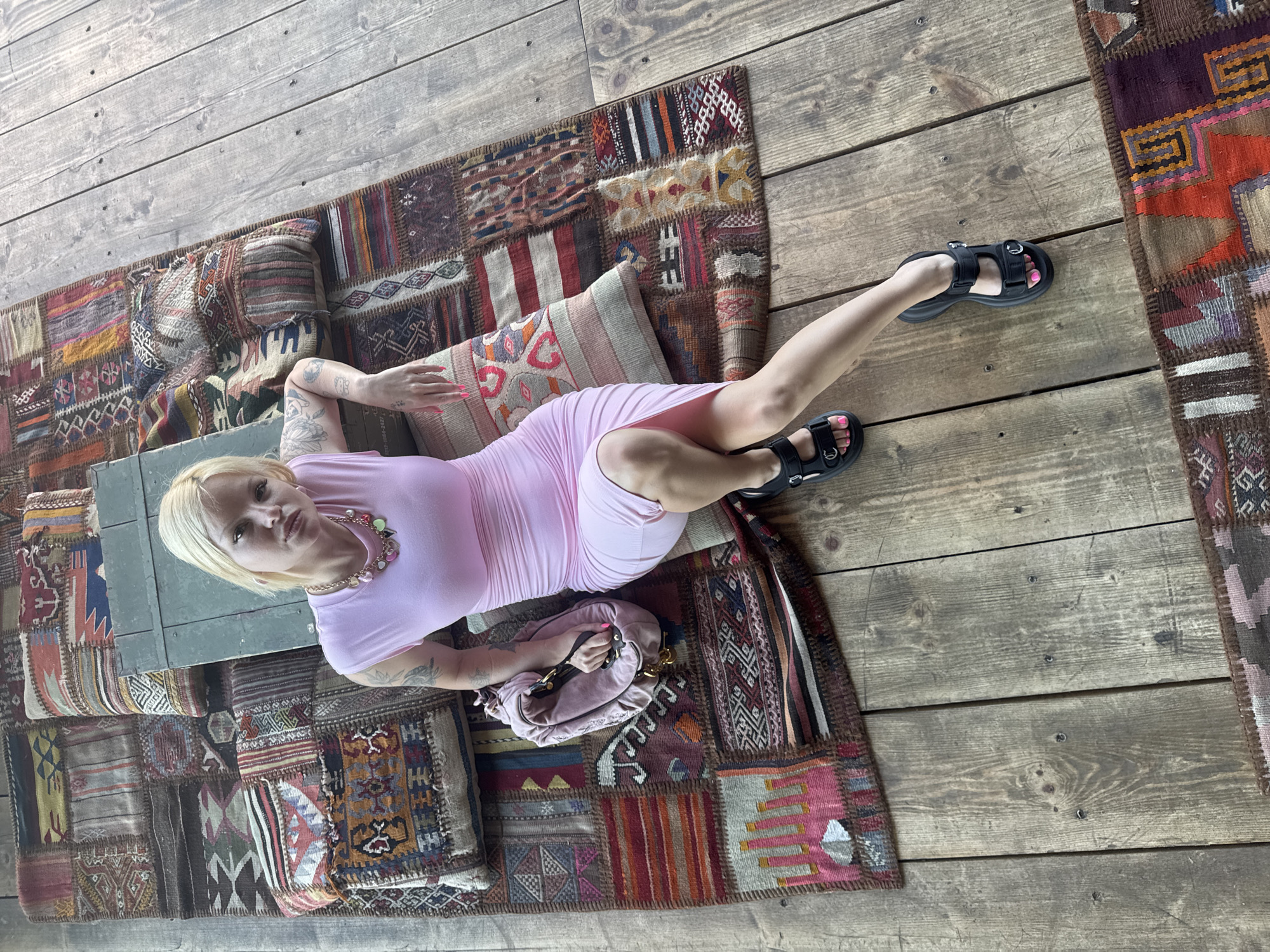 A woman with blonde hair and tattoos on her arms sits on a patchwork rug placed on a wooden deck. She is wearing a pink, ruched dress and black sandals. A pink handbag rests beside her. A vintage green metal chest is positioned behind her. The scene is outdoors, likely on a deck or patio, with visible wooden planks and a colorful, patterned rug. The setting appears to be a casual outdoor space. There is no specific indication of the city of Belgrade, Serbia, within the image itself.