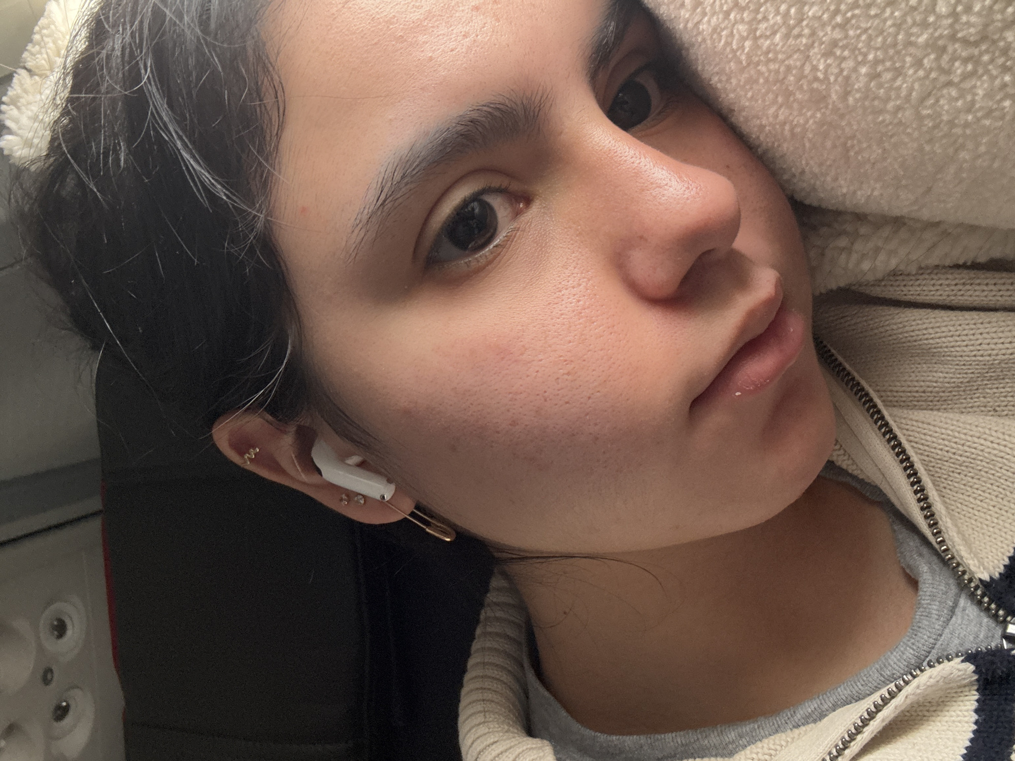 A close-up, angled shot captures a young woman's face and upper torso. She has dark hair, brown eyes, and a fair complexion. In her left ear, she wears an earbud, multiple earrings, and a gold safety pin. She is wearing a zipped sweater layered over a gray shirt. Her face rests against a light-colored fluffy material. The background features a dark surface with a white circular object with dark dots. The location is undetermined; Maramec, United States is not discernible from the image.