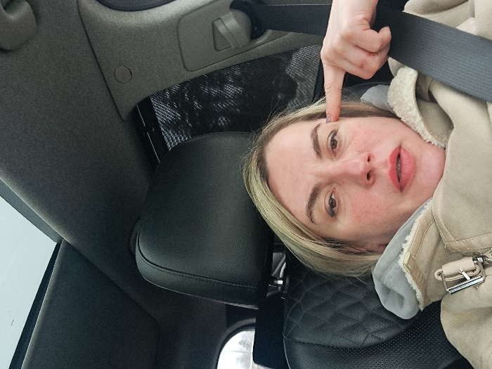 The image shows an adult female seated in the driver's seat of a vehicle, in Obukhovo, Russia. She has blonde hair and is wearing a beige winter coat with a faux fur lining and a visible metallic buckle. A black seatbelt is positioned across her chest and lap. Her right hand is raised, with the index finger pointing towards her right temple. She is looking directly at the camera. The vehicle's interior includes a dark headliner, a black leather seat with a quilted pattern on the seatback, and a panoramic sunroof overhead revealing an overcast sky. A dark mesh sunshade is visible on the passenger side window, partially obscuring blurred outdoor vegetation.
