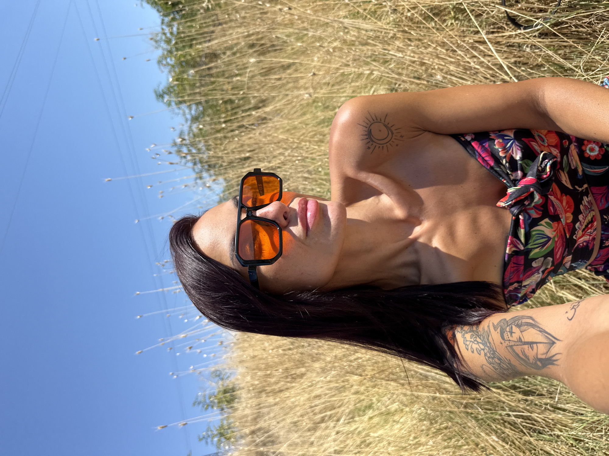 A medium shot shows a woman from the chest up, posed in a field of dry grass under a clear blue sky with power lines visible in the distance. She has long, dark hair, is wearing large, black-rimmed sunglasses with orange lenses, and a strapless bikini top with a floral pattern. The woman has tattoos on her left arm and right shoulder. The scene appears to be outdoors in Pleven, Bulgaria.