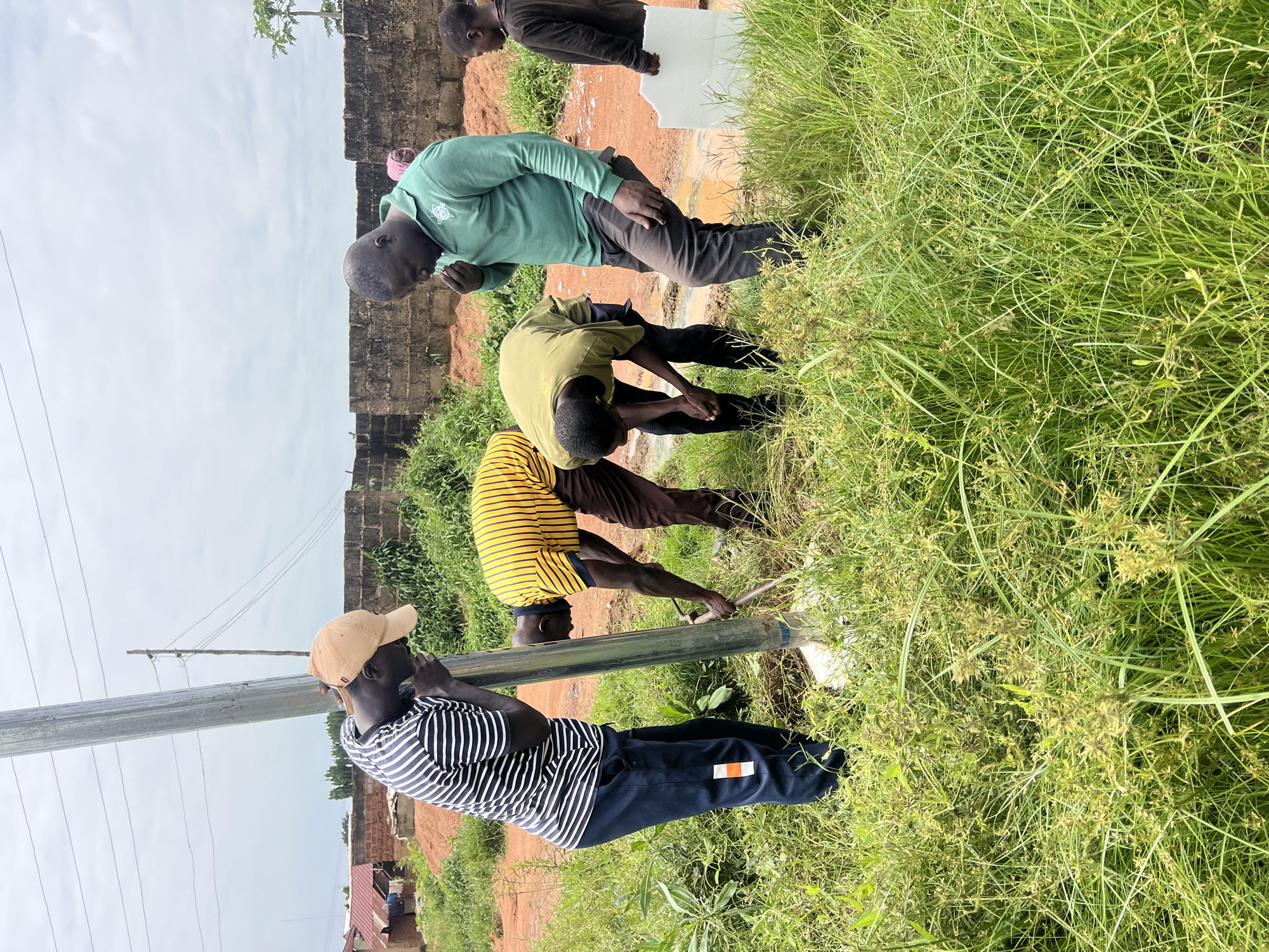 In Jos, Nigeria, four men are engaged in the installation of a metal pole. The setting appears to be an outdoor, semi-rural area with sparse vegetation, including tall grasses and some bushes. In the background, there are brick structures and a cloudy sky. The men are dressed in casual attire. One man in a striped t-shirt and a tan baseball cap leans against the pole, seemingly observing or assisting. Another man, wearing a yellow and black striped t-shirt, is bent over, appearing to work at the base of the pole with a tool. A third man in a green polo shirt stands nearby, looking down at the ground. A fourth individual, partially visible and wearing a dark top, stands to the right, holding a flat white object. The ground around the pole is disturbed, suggesting recent digging or preparation.