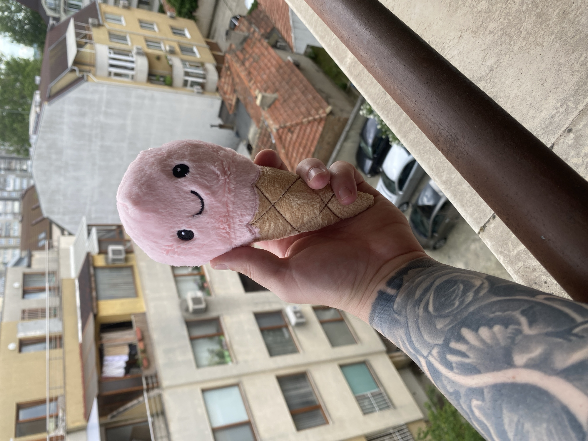 A person with tattooed arm is holding a plush toy resembling an ice cream cone. The toy has a pink, round top with a simple smiling face and a cone-shaped bottom with a brown checkered pattern. The person is standing on a balcony with a brown metal railing visible in the foreground. The background shows a cityscape with apartment buildings of varying heights and architectural styles, typical of urban areas. A building with red roof tiles is also visible. The image may have been taken in Varna, Bulgaria, but without additional information about specific landmarks, a precise determination of the location is not possible.