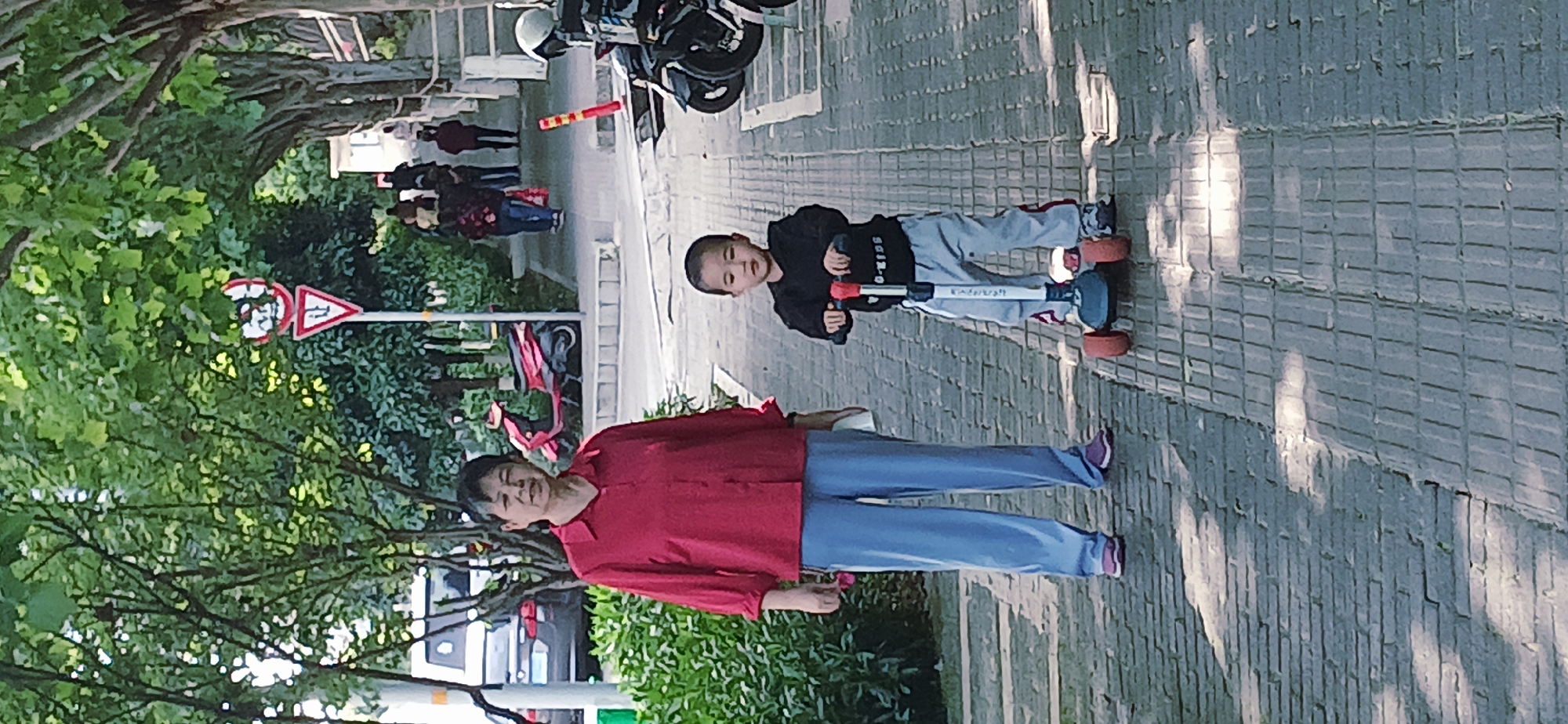 This image captures a vibrant outdoor scene on a paved sidewalk, likely in Jiulongpo District, China. The primary subjects are an adult woman and a young boy, both appearing cheerful.
The woman, positioned on the left side of the frame, is an older adult with short, dark hair and a pleasant smile. She wears a bright red long-sleeved shirt, blue pants, and dark-colored sneakers with white soles. To her right stands a young boy, also smiling towards the viewer. He has short, dark hair and is dressed in a black long-sleeved shirt with white text, possibly "SGIZ-O," and grey sweatpants with red stripes, paired with dark sneakers. The boy is standing on a three-wheeled scooter with a light blue base, a red handlebar, and orange-red wheels.
The setting is a tree-lined street. Lush green foliage from numerous trees and bushes frames the left side of the image and extends into the background. A paved road runs parallel to the sidewalk, with several other people visible in the distance, some walking, and others indistinct. A parked black motorcycle or scooter is seen further back on the sidewalk. Amidst the greenery, two traffic signs are noticeable: a red circular sign with a white bar (likely "No Entry" or "No Vehicles") and a red-bordered triangular warning sign.
The activity suggests a casual outing, possibly a leisure walk for the woman while the boy enjoys riding his scooter. The bright lighting, with shadows cast on the patterned grey sidewalk, indicates it is daytime, likely a sunny and pleasant day. Both individuals exhibit contented expressions, suggesting a relaxed and happy atmosphere.