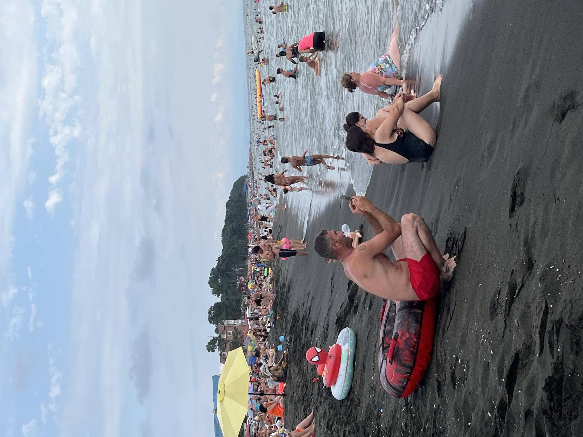 The image depicts a crowded beach in Ureki, Georgia, known for its black magnetic sand. In the foreground, a man in red swim trunks is crouching on the dark sand, holding a smartphone and seemingly taking a photo or video. Beside him, a red inflatable tube with a Spiderman design is partially visible. Further down the beach, several people are sitting on the sand, including a woman in a black swimsuit and another in a floral swimsuit. The middle ground of the image shows a multitude of people wading and swimming in the sea, with some engaging in recreational activities. In the background, a yellow umbrella provides shade for beachgoers, and buildings and lush greenery line the coast. The sky is overcast with scattered clouds. The overall scene captures a lively summer day at a popular seaside resort.