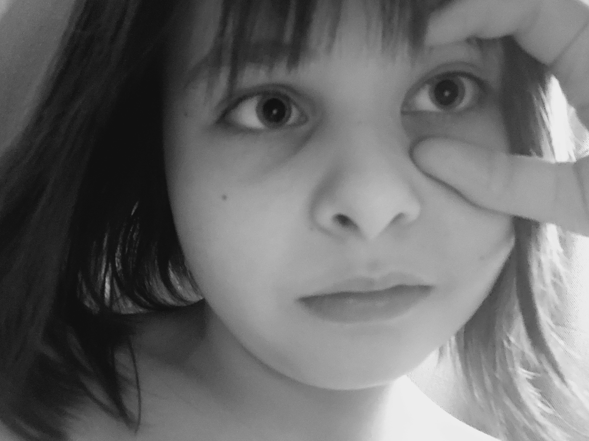 A close-up, grayscale image shows a young person with dark hair and bangs. Their face is the primary focus. The person is looking directly at the viewer with wide eyes. A hand with fingers spread is partially obscuring the right eye. The skin tone appears light. No location information is discernible in the image, so it's impossible to determine if it was taken in Izhevsk, Russia or any other specific location.