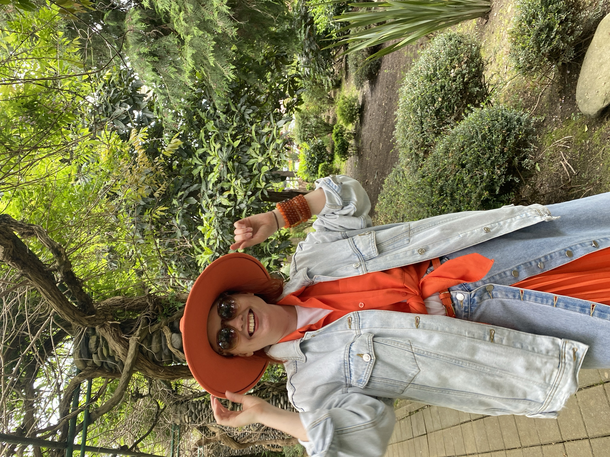 A person stands amidst lush greenery, likely in a garden or park setting. The individual wears an orange hat and sunglasses and dons a denim jacket over an orange shirt, paired with light blue jeans. The background features a variety of trees and bushes, creating a natural environment. The location may be Sochi, Russia. The image appears to have been rotated.