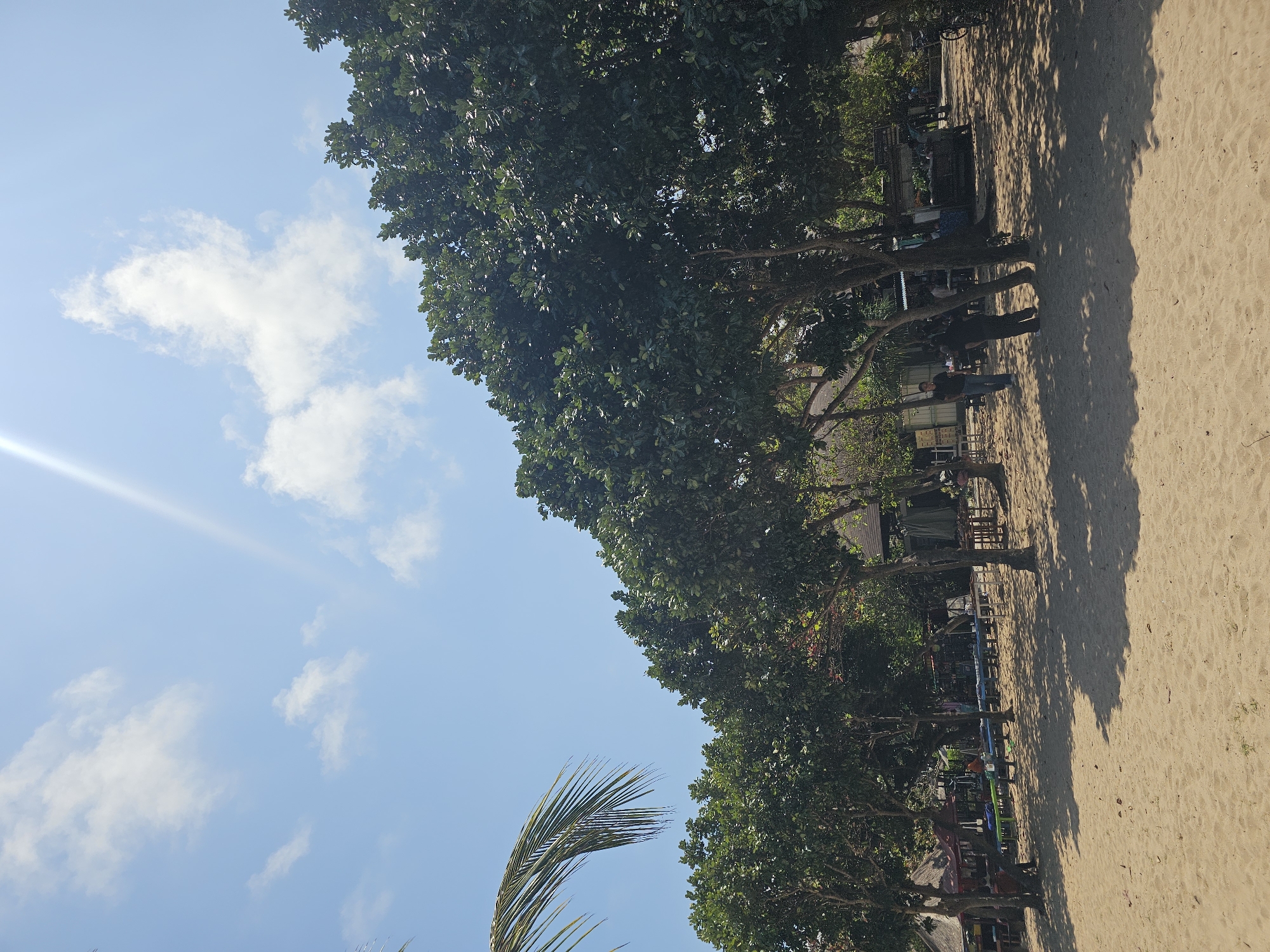 This is a sunny, daytime, eye-level, outdoor, full shot photograph taken on a sandy beach in Denpasar, Indonesia. In the foreground is a wide expanse of light brown sand, with gentle undulations and shadows cast by trees. A frond of a palm tree extends from the upper left corner. Behind the sand, a row of dense, green-leafed trees runs diagonally across the midground. These trees are casting dappled shadows on the sand. Beyond the trees, a few small structures with thatched roofs are visible, along with some tables and chairs. There are several people scattered throughout the midground and background, some appearing to be walking or standing near the structures, while others are seated at tables. The sky above is a clear, bright blue with a few wispy white clouds and a lens flare originating from the upper right corner. The overall scene depicts a lively beach area on a bright, clear day.