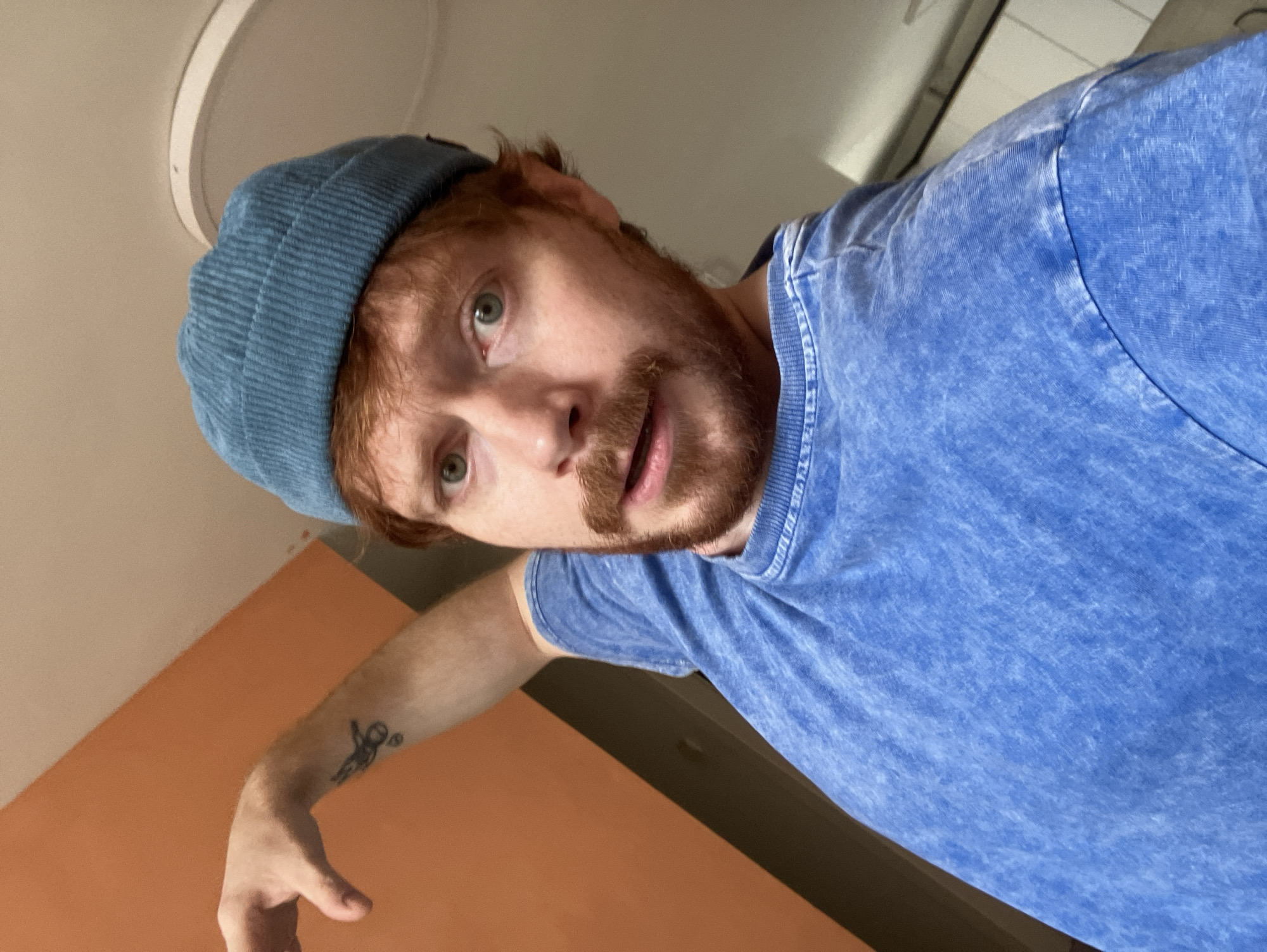 A man with red hair and a beard is wearing a blue t-shirt and a blue beanie. He has a tattoo of a small figure on his left forearm. The background includes orange and white walls, and a ceiling light fixture. There is no information to establish that this photo was taken in Tel-Aviv, Israel.