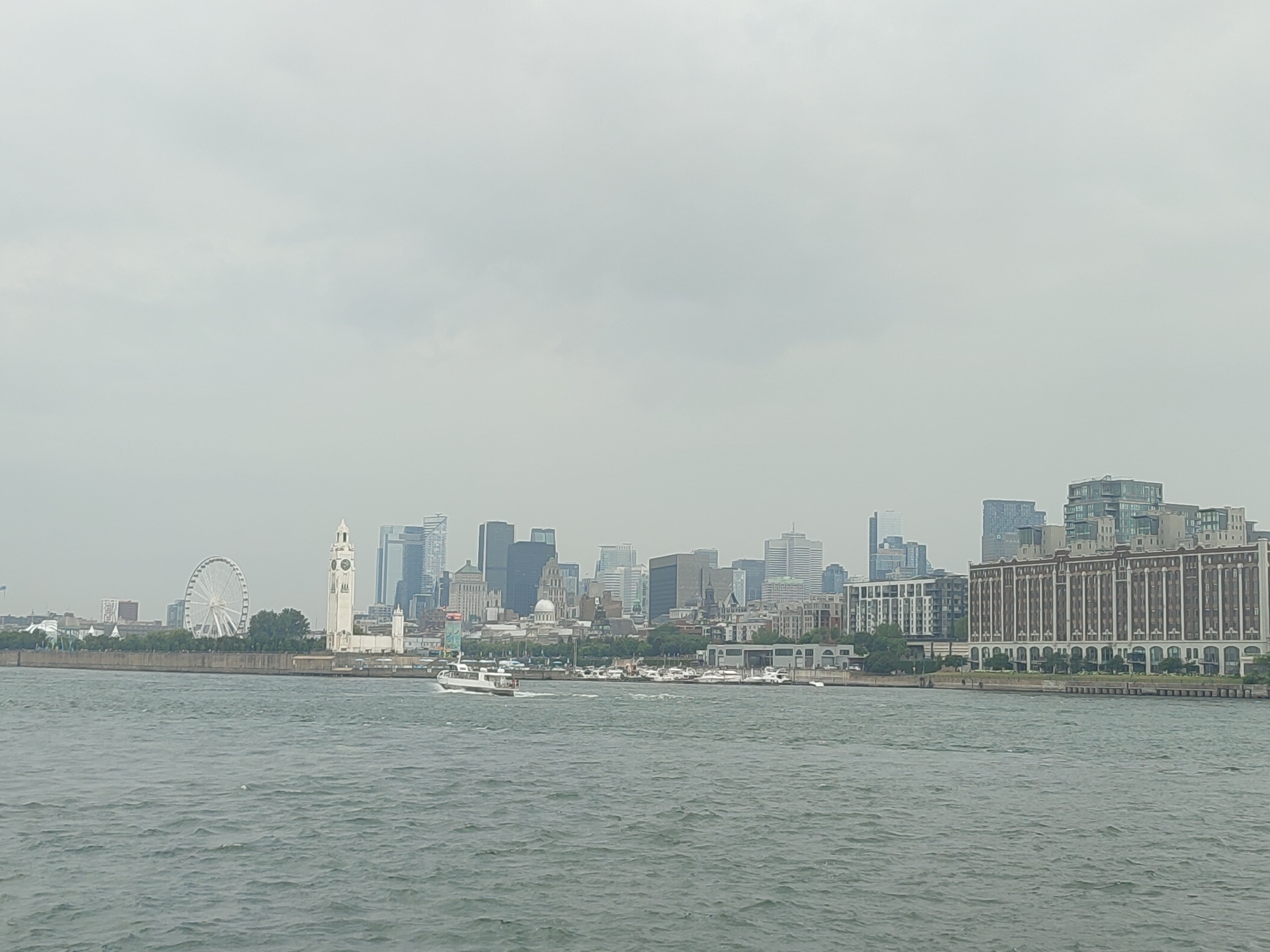 The image shows a panoramic view of the Montreal skyline from across the water. In the foreground, choppy grey water stretches across the frame, with a small white boat moving from left to right. Along the middle ground, a textured stone embankment lines the waterfront. On the embankment, a Ferris wheel is visible on the left, followed by a tall, white clock tower. Behind these landmarks, a dense collection of modern skyscrapers forms the city's iconic skyline. To the far right, a large, multi-story brick building with numerous windows stands prominently. The sky above is overcast and hazy, casting a diffused light over the entire scene.