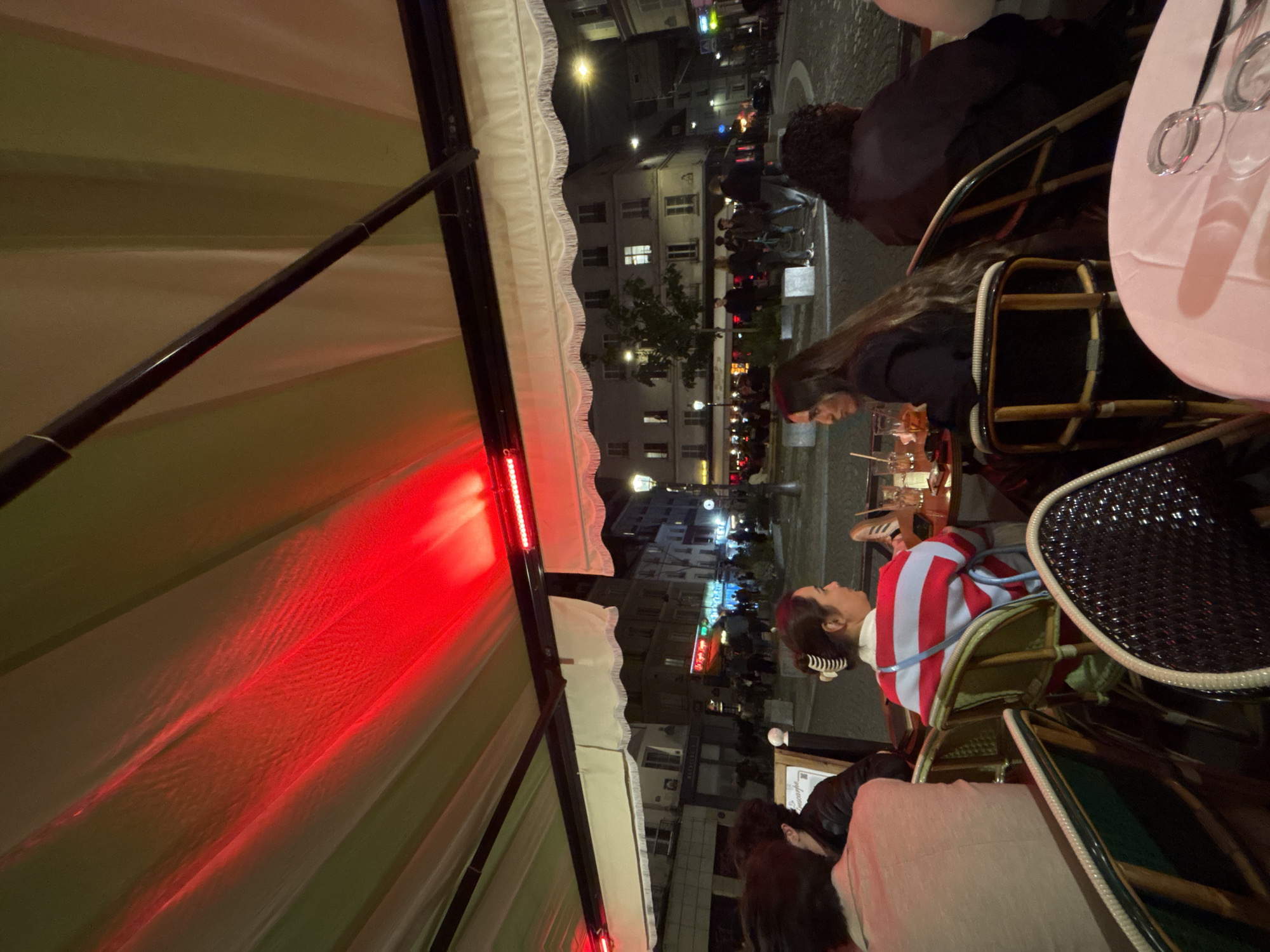 Captured from a slightly angled perspective, the scene unfolds within an outdoor café in Paris, France. Underneath a striped canopy lit by red lights, people are seated at tables. One person is wearing a striped shirt, while another is wearing a red beret. Clear glasses and other items are on a pink table, suggesting diners are engaged in conversation and dining. The café is illuminated by streetlights, and in the background, buildings line the street, hinting at a bustling urban environment. Pedestrians can be seen walking along the sidewalk beyond the café's perimeter.