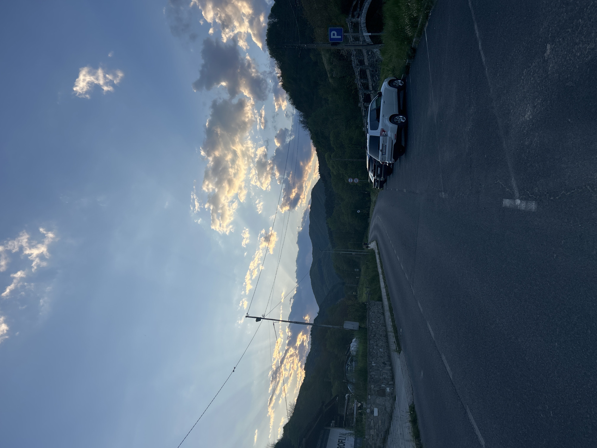 The image captures a scenic view near Zlatograd, Bulgaria. In the foreground, a paved road stretches out of view with a line of parked cars on the right side, including a silver SUV. Adjacent to the road is a low stone wall with some greenery. Behind the cars, a steep, tree-covered hillside rises, partially obscuring a parking sign. The background features a mountainous landscape and a sky filled with scattered, sunlit clouds. A utility pole and wires are visible against the sky, adding to the rural atmosphere.