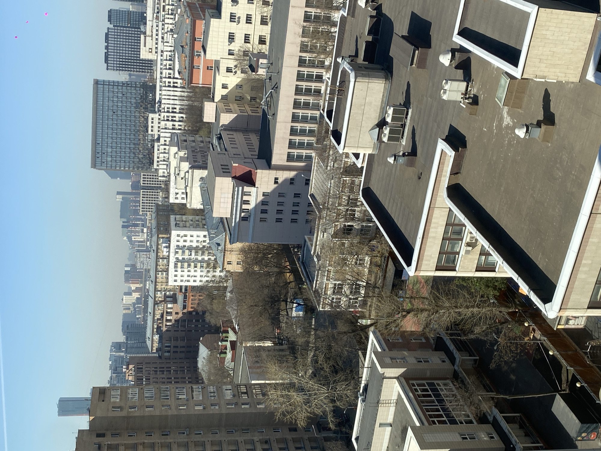 The image depicts a cityscape of Moscow, Russia, viewed from an elevated position, possibly a high-rise building. The scene is dominated by a variety of buildings, ranging from older, multi-story structures with many windows to more modern, taller skyscrapers. The buildings are densely packed. There are also scattered trees, some bare, which suggest it may be early spring or late autumn. In the foreground, the roofs of buildings are visible, with various HVAC units and other rooftop equipment. The sky is a clear blue. There are some small pink objects, possibly balloons, floating in the sky.