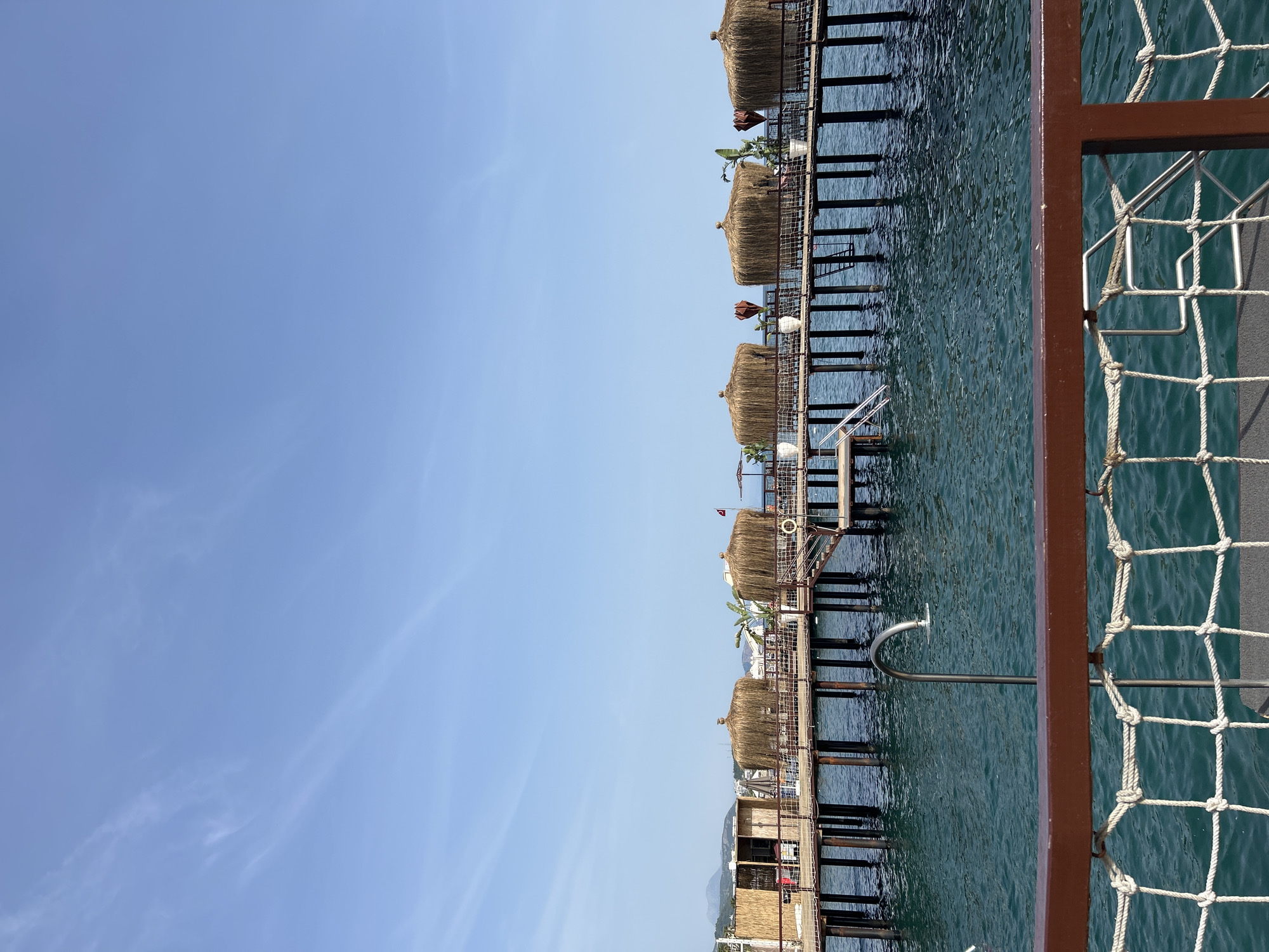 This image depicts a series of thatched-roof bungalows or cabanas extending out over the turquoise water. They are connected by wooden walkways. In the foreground, a brown metal railing with white netting is visible. The sky is clear and blue. The location is Alanya, Turkey.