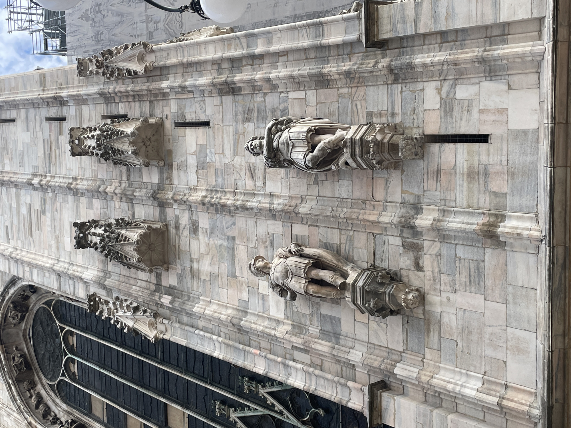 The image shows a close-up of the exterior of the Duomo di Milano, a large Gothic cathedral in Milan, Italy. The building is made of white marble blocks. Two statues of figures, likely saints or important religious figures, are affixed to the facade. Above each statue is an elaborate stone corbel, while a vent is located to the right of the statues. A portion of a large stained-glass window is also visible on the lower left. Architectural details such as molding and ornamentation are present along the surface. The background is a cloudy sky.