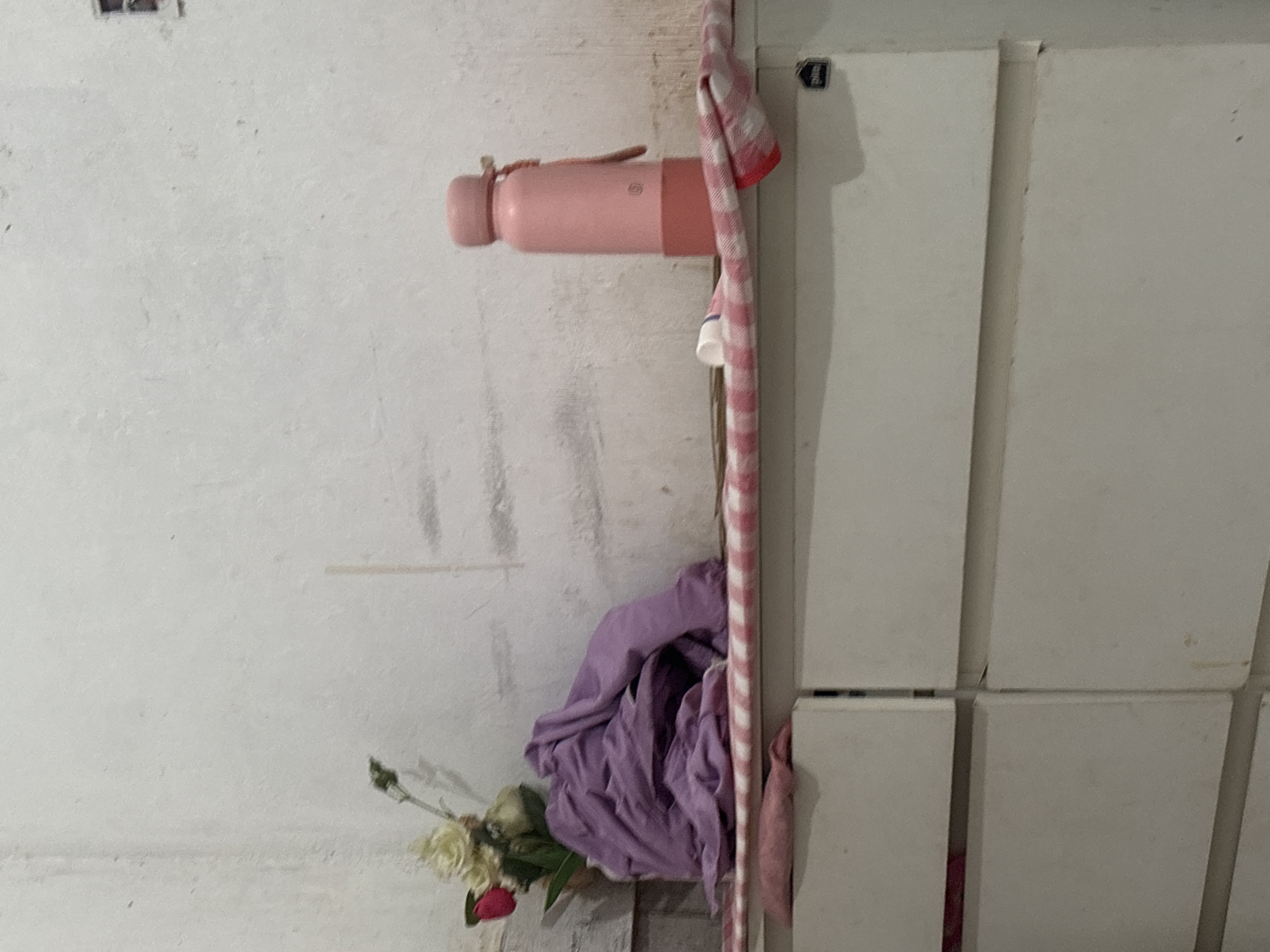 The image depicts a close-up, eye-level shot of a white, textured wall with visible smudges and streaks, suggesting it is in a lived-in space, possibly in North Jakarta, Indonesia. On a shelf or surface in the foreground, a pink water bottle with a matching pink lid stands upright. Beside the water bottle, a small white cap from a container is visible. A pink and white checkered cloth is draped over the surface, extending slightly over the edge. To the left of the water bottle, a bundle of purple fabric is piled up, and behind it, a bouquet of artificial flowers with white and pink blossoms is placed. Beneath the shelf, the top edges of white drawers are visible. The overall impression is of a cluttered shelf in a domestic setting.