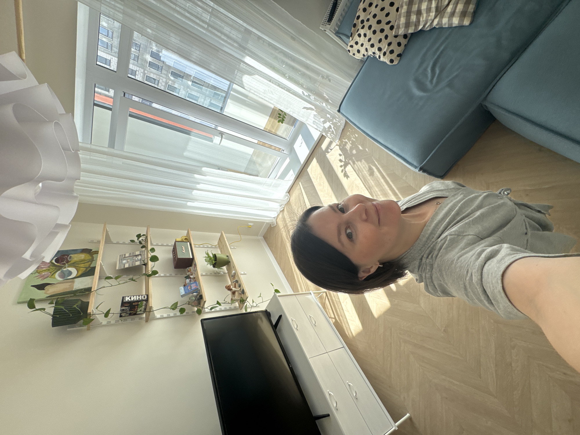 A selfie shot taken indoors in Izhevsk, Russia. A person with dark hair, wearing a gray sweatshirt, holds the camera. Behind the person is a living room area. A light blue sofa with decorative pillows and a throw blanket is located in the upper right corner of the frame. A window with white curtains stretches along the top side of the image, providing natural light. To the left, wall-mounted wooden shelves display various decorative items, including a painting, books, and a speaker. A television screen is located in the lower-left corner. A white cabinet with drawers is placed next to the television. The floor has a light-colored parquet pattern. The lighting appears natural and bright.