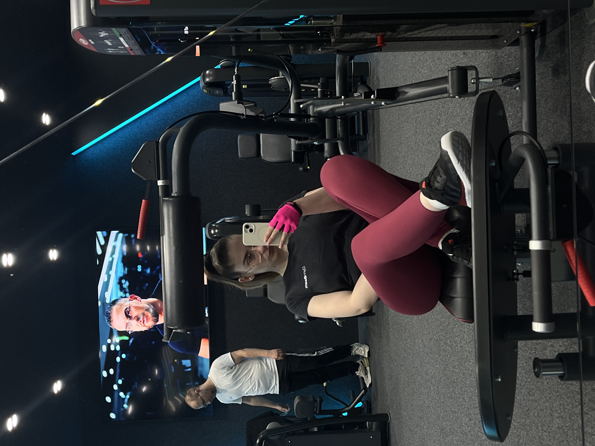A young woman wearing a black t-shirt, maroon leggings, and pink gloves is taking a selfie with her smartphone while sitting on a leg press machine at a gym in an unknown city. A man in a white t-shirt and black pants is seen in the background. A television screen shows a close-up of a man with glasses. The gym features black walls and blue accent lighting.