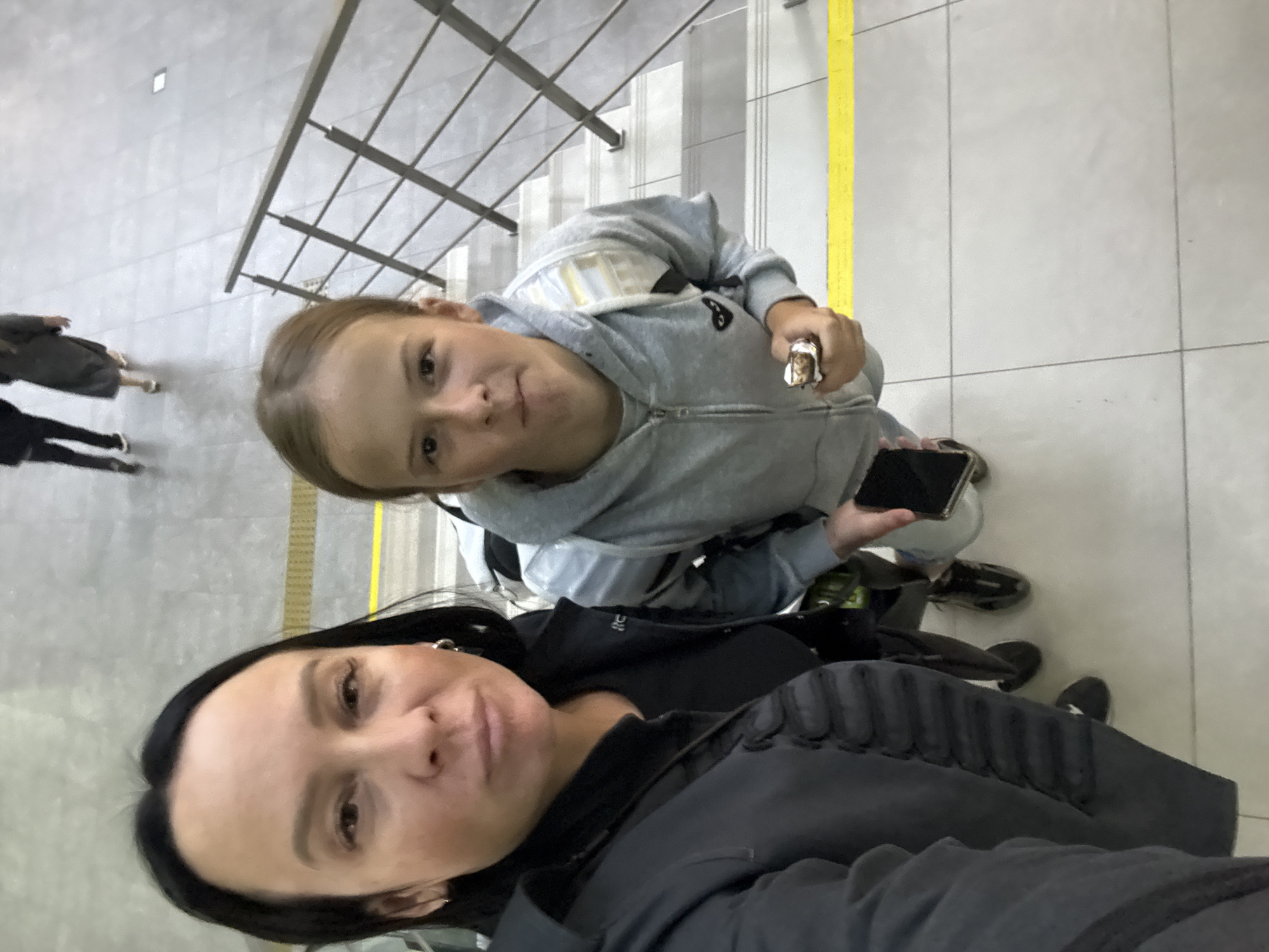 In Bolshoye Savino, Russia, at an airport terminal, a woman and a young girl are taking a selfie on a staircase. The woman, with dark hair pulled back, wears a dark jacket over a black shirt and silver hoop earrings. The girl, with light brown hair, wears a grey hooded sweatshirt with a heart-shaped logo and a backpack. She holds a phone and a snack bar. Behind them, two individuals walk away, their figures blurred. The staircase is made of concrete and metal, with yellow safety markings on the steps.