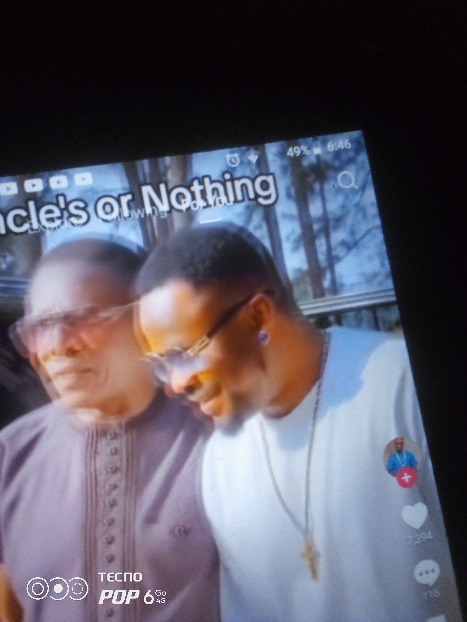 Two men are shown in a close-up shot. The man on the left, wearing glasses and a brown shirt, is partially out of focus. The man on the right, wearing a white shirt and glasses, is in sharper focus. He has a purple earring in his left ear and a cross pendant on a chain around his neck. There is a profile picture with a plus sign next to it on the right side of his shirt, followed by a heart icon with "7,394" below it and a speech bubble icon with "116" below it. Text at the top of the image reads "Uncle's or Nothing" and the bottom of the image has "TECNO POP 6 Go 4G" and other logos. The background appears to be outdoors with trees and a railing. The image is a screenshot of a video or social media post.