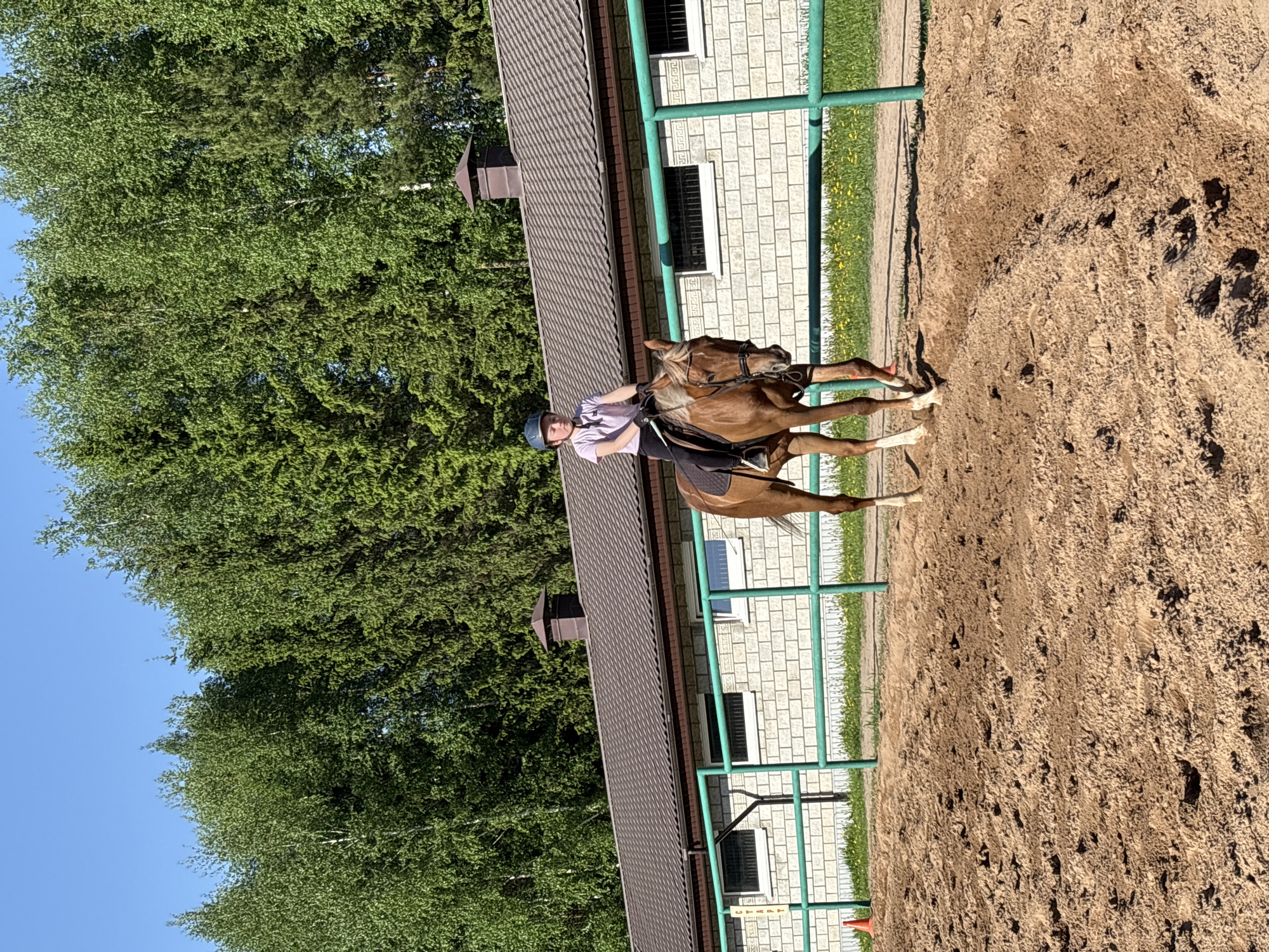 A person wearing a helmet and light-colored shirt is riding a brown horse in an outdoor riding arena. The arena has a sandy surface and is enclosed by a green fence. A brick building with a dark roof runs alongside the arena. Trees are visible in the background under a blue sky. The location appears to be in Русский Вожой, Russia.