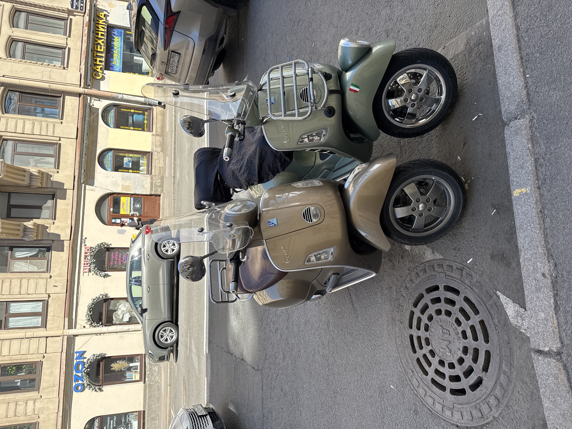 This image depicts a sunny urban street scene on Razyezzhaya Ulitsa (Разъезжая улица), specifically near building number 21 as indicated by a street sign, in Saint Petersburg, Russia.
In the immediate foreground, two vintage-style Vespa scooters are parked on an asphalt surface. The left scooter is a metallic brown/gold color with a brown leather seat, displaying the "Vespa" script and a Piaggio "P" emblem. The right scooter is olive green, featuring a black protective cover over its seat, a chrome front rack, "Vespa" script, a Piaggio "P" emblem, and a small Italian flag sticker on its front fender. Both scooters have clear windshields, chrome rearview mirrors, and multi-spoke chrome wheels. A circular metal manhole cover with "WT C250" and Cyrillic text is visible on the asphalt in the lower-left corner. The ground around the scooters and near the grey concrete curb is littered with numerous white cigarette butts.
In the mid-ground, a grey Audi Q3 SUV is parked on the street. Behind it, partially visible on the right, is the rear section of a silver Lexus RX SUV, displaying a Russian license plate "B693TP 198," which is registered to Saint Petersburg. Another car, a silver sedan with a license plate "042YP 98" (also Saint Petersburg region), is partially visible in the far left foreground.
The background features a multi-story, light-colored building with numerous brown-framed windows on its upper floors. The ground floor houses several businesses. From left to right, visible businesses include "Ozon" (an online retailer) with blue lettering and white floral decorations around its arched windows, "ЦВЕТЫ" (Flowers) with pink lettering and similar floral decorations, and "САНТЕХНИКА" (Plumbing) with yellow and blue signage. A brown door labeled "ВХОД БАРБЕРШОП" (Barbershop Entrance) is also visible. A person in dark clothing is partially visible walking away from the barbershop entrance on the right side of the street. The scene is brightly lit by direct sunlight, casting distinct shadows.