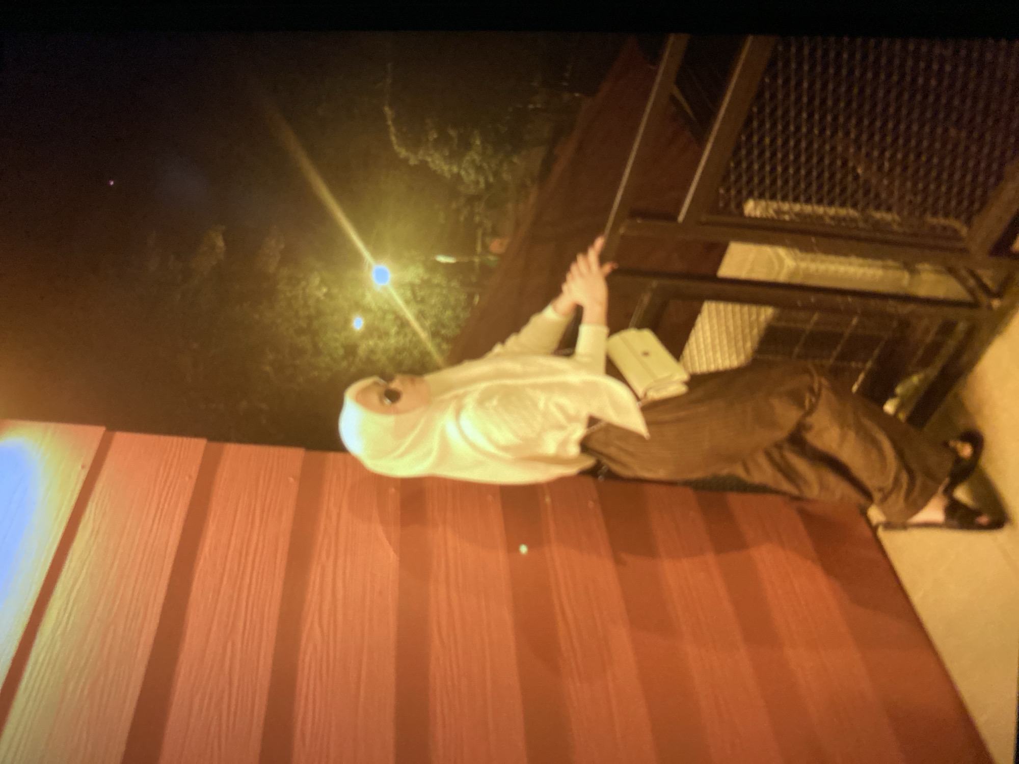 A young woman wearing a white hijab, sunglasses, a white long-sleeved shirt, and brown striped pants stands on a balcony at night. She is leaning on a dark railing, looking out into the distance. The background shows trees and a bright yellow street light. The building behind her is made of red wooden planks. The scene appears to be outdoors, possibly in a residential area or a cafe in Ta', Indonesia.