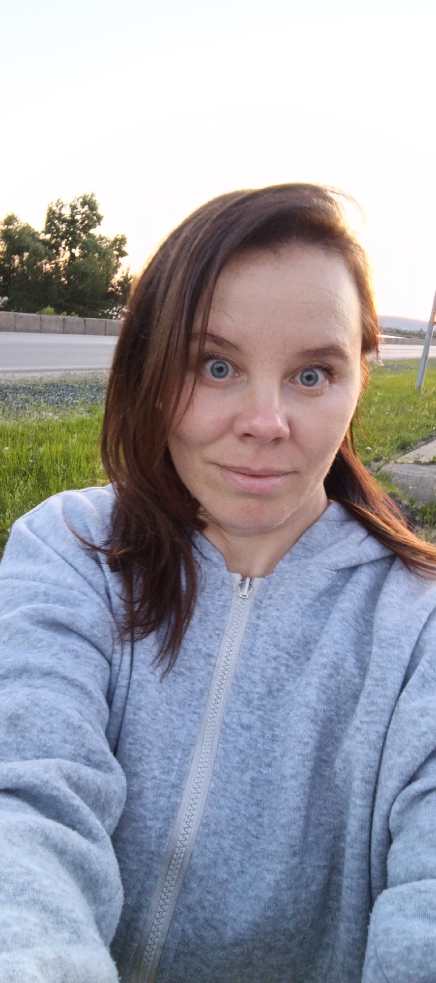 A woman with reddish-brown hair and blue eyes is taking a selfie outdoors in Chaikovsky, Russia. She is wearing a light gray zippered hoodie. The background shows a grassy area with a road and some trees. The sky is bright, suggesting it is daytime, possibly late afternoon or early evening due to the warm lighting.