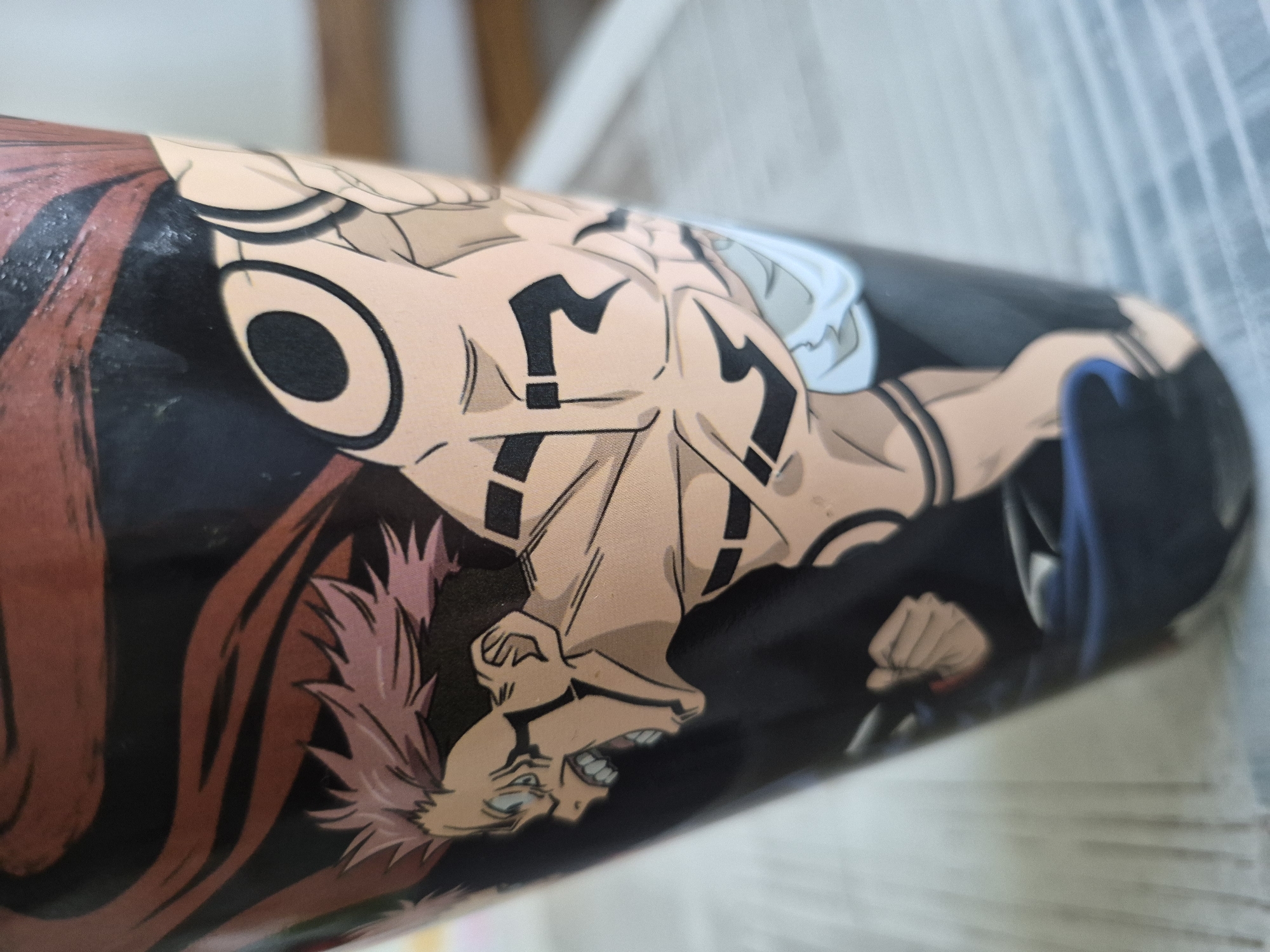 The image is a close-up shot of a cylindrical object, possibly a cup or a container, with a printed illustration of an anime character on it. The character has spiky pink hair, an aggressive facial expression with teeth bared, and visible tattoos on its chest and arms. The tattoos are black and geometric in design. The background of the illustration is black, with some dark red flowing elements above the character's head. The object is placed on a surface that appears to be a table with a textured, light-colored top, and a blurry background suggests an indoor setting. There is no indication of a city or specific location.