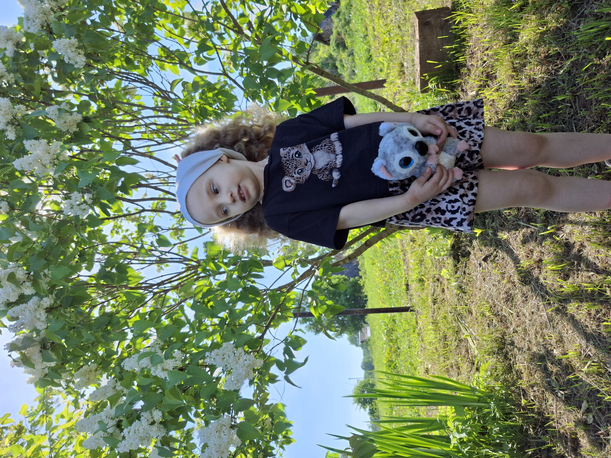 A young girl stands outdoors, likely in a garden or park-like setting. She is wearing a dark t-shirt with a leopard print on the front, a skirt with a similar print, and a light-colored headband. She is holding a stuffed animal, possibly a koala, in her hands.
The background includes a flowering bush with white blossoms, possibly lilac, and green grass. Part of a wooden structure is visible in the upper right. There are also green reeds visible on the edge of the image, along with a fence. The sky is blue and sunny. Given the location of Nizhnyaya Gar', Russia, the scene is likely taking place there in the spring or summer.