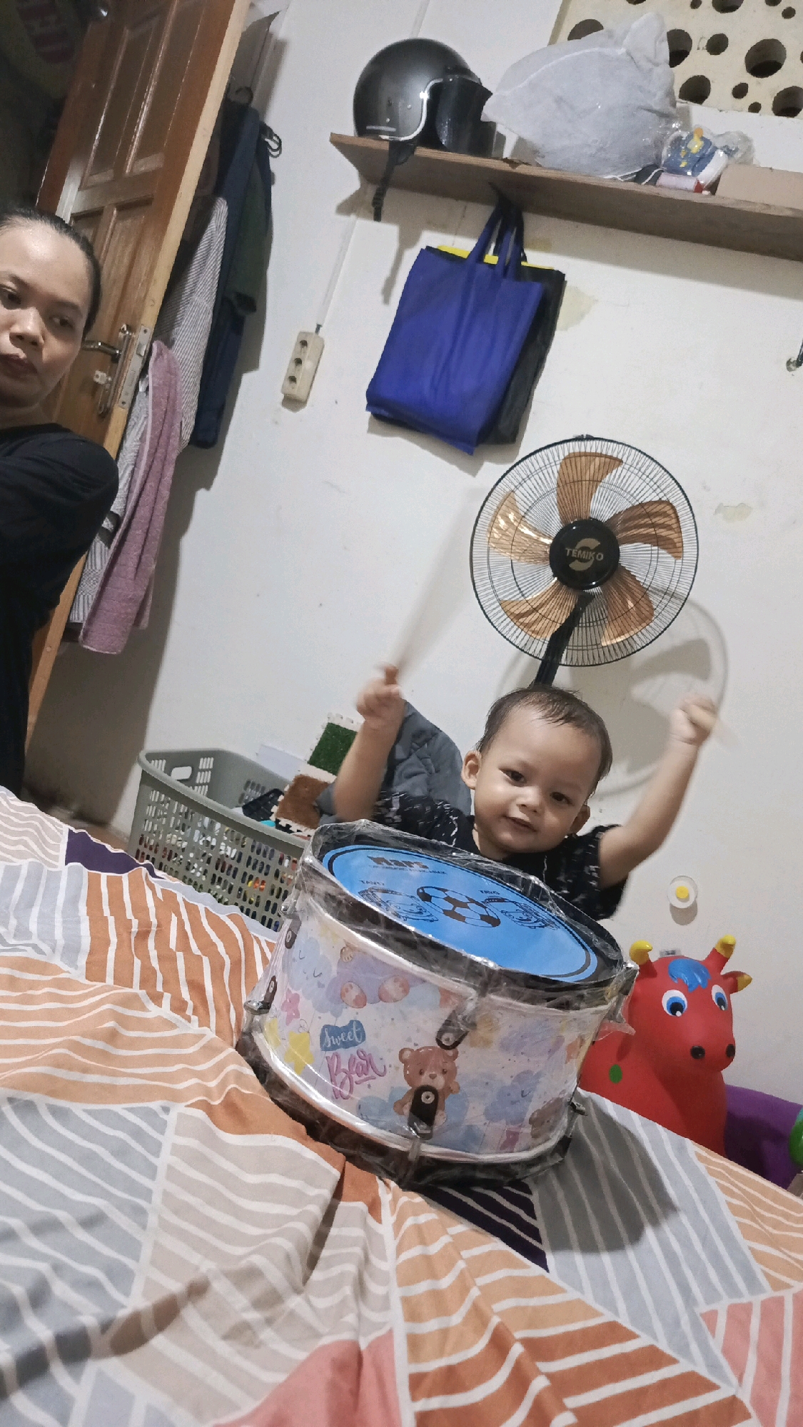 In a room in Pekauman Kulon, Indonesia, a young child is seen sitting on a bed, enthusiastically playing a drum. The child, wearing a black shirt, has their arms raised and appears to be hitting the drum with drumsticks, their face beaming with joy. The drum itself is decorated with a colorful cartoon pattern featuring bears and clouds, and it is wrapped in plastic. To the right of the child, a red toy horse with blue horns and big blue eyes is visible. The bed is covered with a comforter featuring a geometric pattern of horizontal stripes in various colors including orange, gray, white, and pink. In the background, a black oscillating fan is mounted on the wall above the child. To the left of the fan, a wooden shelf holds a black helmet and other items. Further to the left, a woman is partially visible, her face turned towards the camera. The room also contains a grey laundry basket filled with clothes and a wooden door. The walls are white and appear slightly textured.