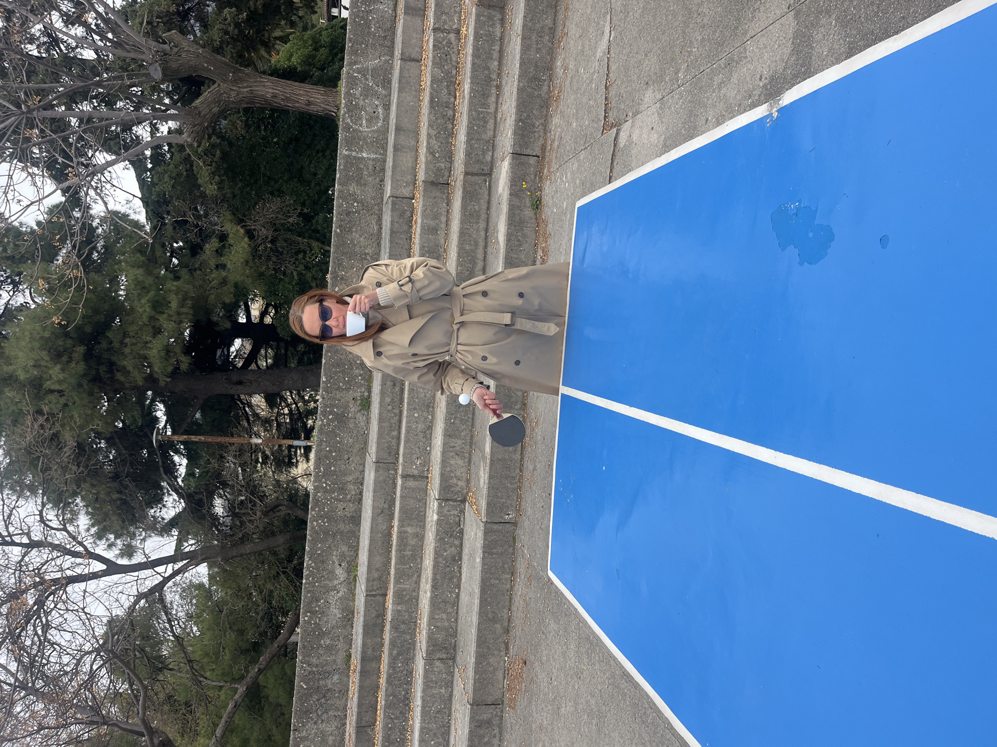 A woman in a trench coat stands in front of a blue ping pong table outdoors in Budva, Montenegro. She holds a table tennis paddle in her right hand and a white cup in her left. She is wearing sunglasses and standing in front of concrete steps. Trees are visible in the background.