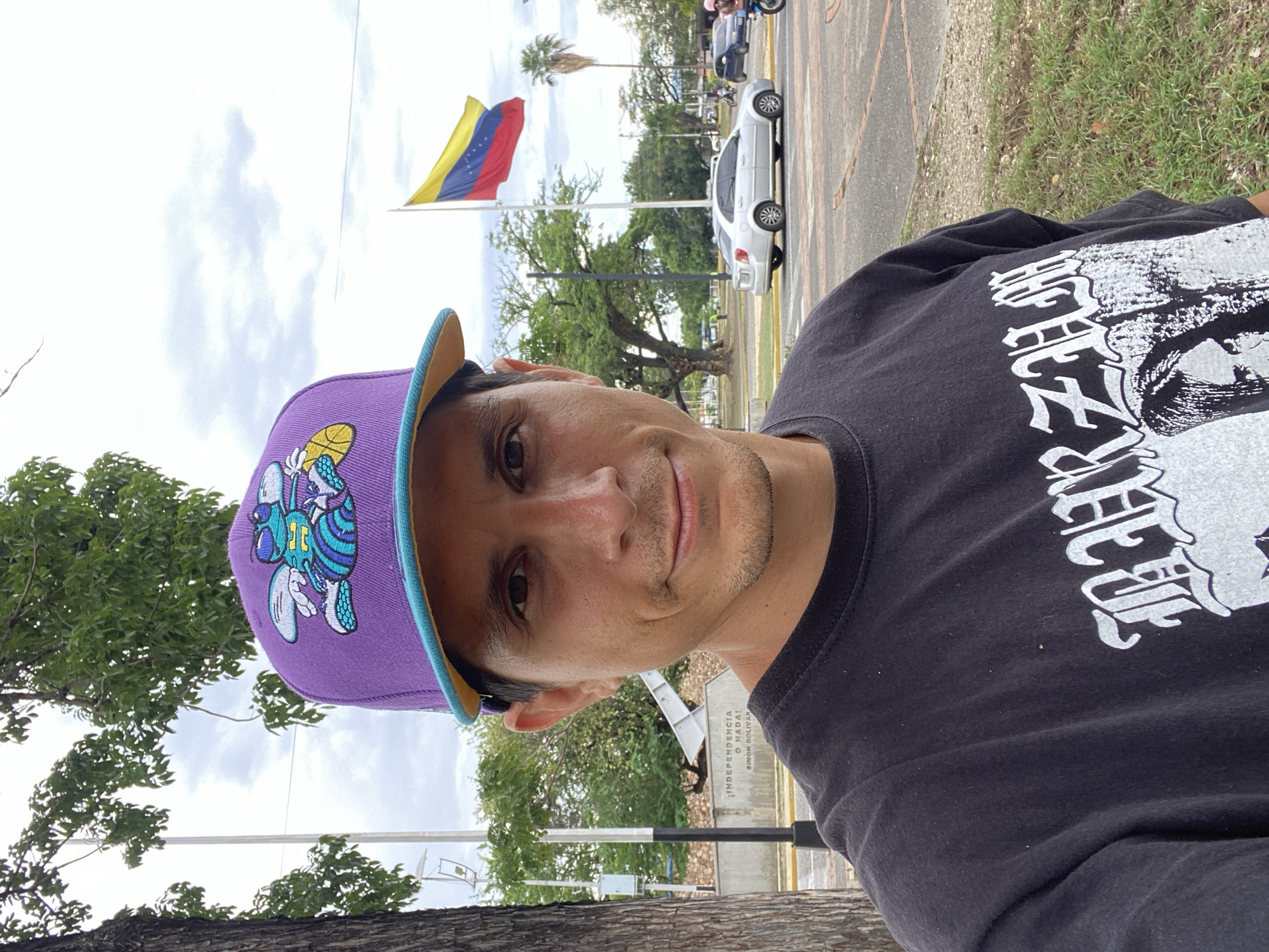 A close-up outdoor selfie, rotated 90 degrees to the left, captures a man against a vibrant background featuring national symbols and a street scene in San Carlos, Venezuela.
The man, positioned in the foreground, has short dark hair and light stubble. He wears a purple baseball cap adorned with a colorful embroidered design of a cartoon bee or hornet playing basketball, likely a sports team logo. His dark t-shirt displays a prominent white graphic design with stylized text that appears to be "INDEPENDEZ" or similar, alongside an artistic illustration. He looks directly at the camera with a neutral to slight smile.
In the background, a large Venezuelan flag, featuring horizontal stripes of yellow, blue, and red with an arc of eight white stars on the blue band, flies prominently on a flagpole. A paved street or pathway is visible to the right of the flag, with a white car (possibly a sedan) and another smaller vehicle further down. Lush green grass, various trees, and foliage complete the outdoor setting under a partly cloudy, overcast sky.
A significant detail is a white monument or plaque visible behind the man's right shoulder, inscribed with the patriotic Spanish phrase: "¡INDEPENDENCIA O NADA!" (Independence or Nothing!) followed by "SIMON BOLIVAR." This inscription, along with the Venezuelan flag, firmly establishes the location context. The photograph appears to be taken during the daytime, under natural, diffused lighting conditions.