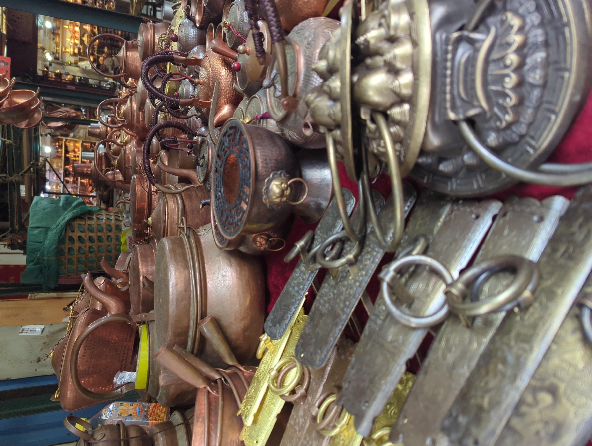 The image depicts a market stall in Gucheng District, China, overflowing with traditional copper and brass cookware and teapots. Numerous teapots of various sizes and designs are stacked and arranged, featuring ornate handles and spouts. Several large copper pots and bowls are also visible, some with intricate patterns. In the foreground, metallic elements with ring handles, possibly part of decorative displays or functional accessories, are laid out. The background hints at a bustling marketplace with shelves of more copper items and stalls. There are no people clearly visible in the frame.