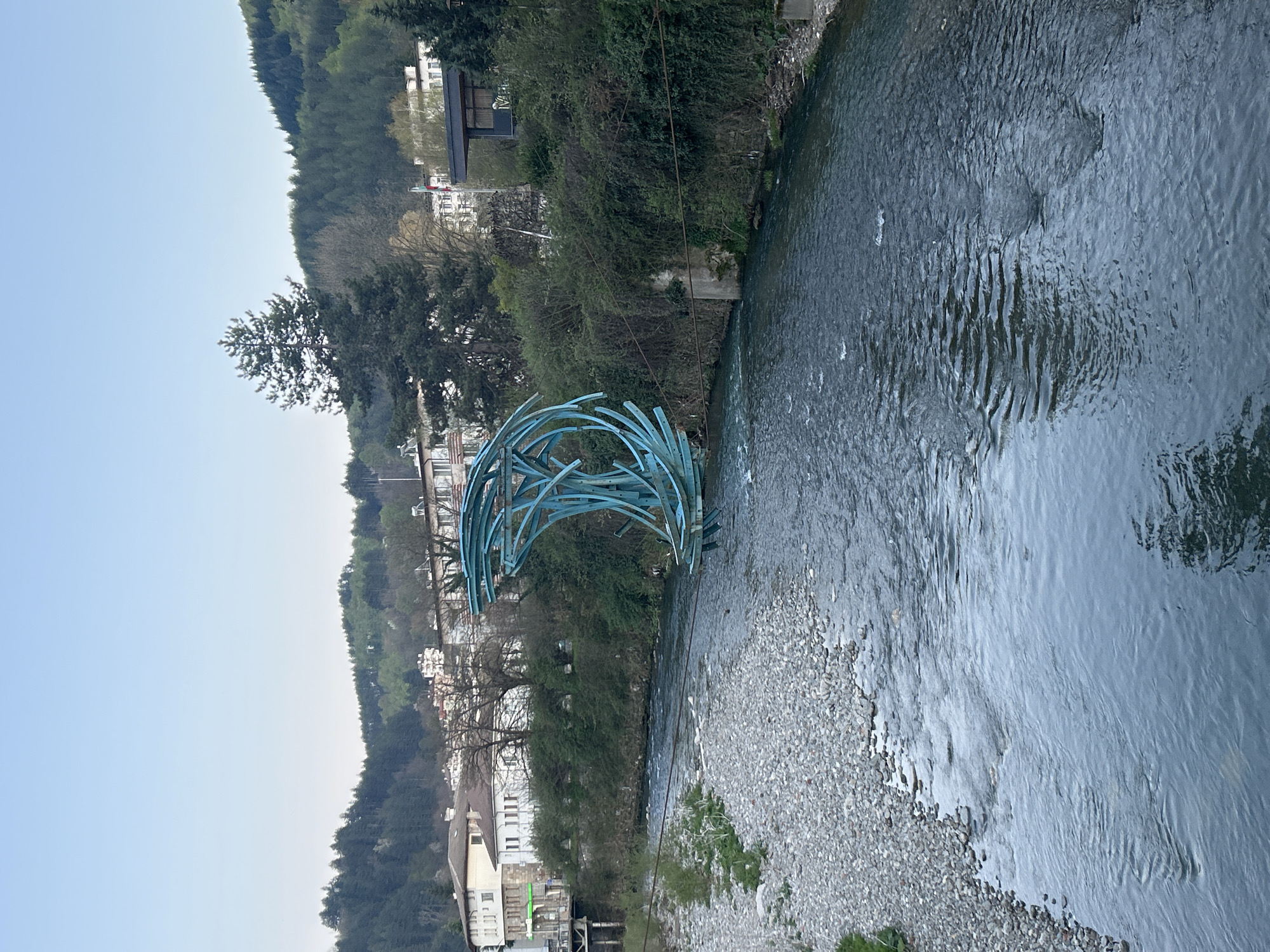 The image displays a panoramic view of a river flowing through a valley, with lush green hills rising on both sides. A distinctive light blue, abstract sculpture, crafted from curved, interconnected pipe-like elements, is prominently featured. It is positioned partially in the river near the left bank, forming an arch or wave-like shape.
The river water shows ripples and reflections, indicating a gentle current, and a pebbly bank is visible along the right side. Both riverbanks and the hills beyond are densely covered with trees and vegetation. Buildings, predominantly white or light-colored, are visible nestled among the trees on both sides of the river, particularly on the left bank. Overhead wires are also visible spanning across the river in the mid-ground.
The scene is captured during daytime under a clear, bright blue sky, suggesting good weather conditions. No specific human activity or visible text is present. The overall impression is one of peaceful natural scenery, augmented by the modern artistic sculpture. This location is consistent with Zlatograd, Bulgaria.