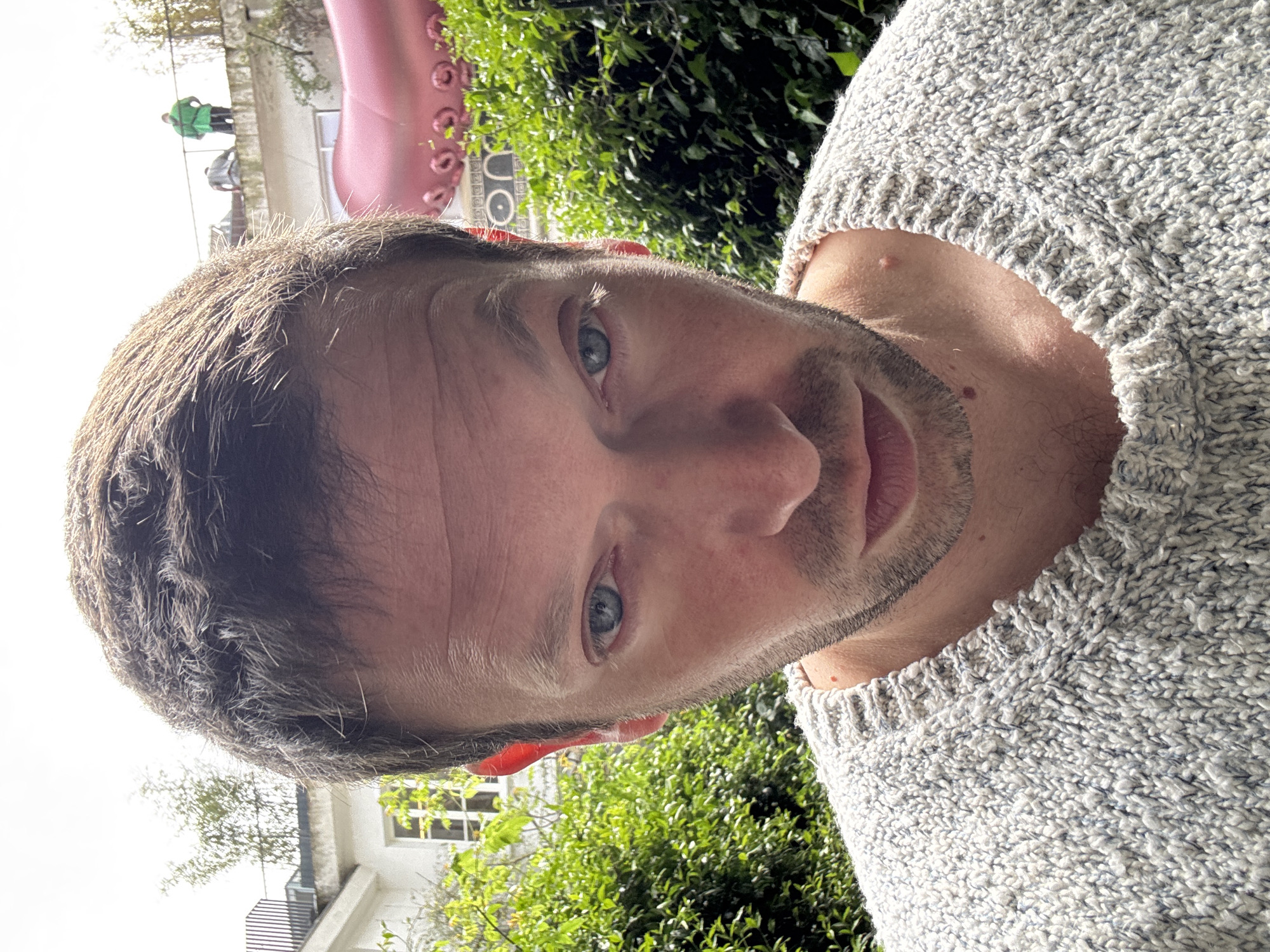 A man with short, light brown hair and blue eyes is posing for a selfie outdoors. He is wearing a grey knit sweater. The background includes green foliage, a building with white walls and windows, and what appears to be part of a large pink inflatable sculpture. In the upper left corner of the image, there are three figures on a roof or elevated platform. Based on the presence of the inflatable sculpture and the architectural style of the building, the location is likely near the Fondazione Prada in Milan, Italy.